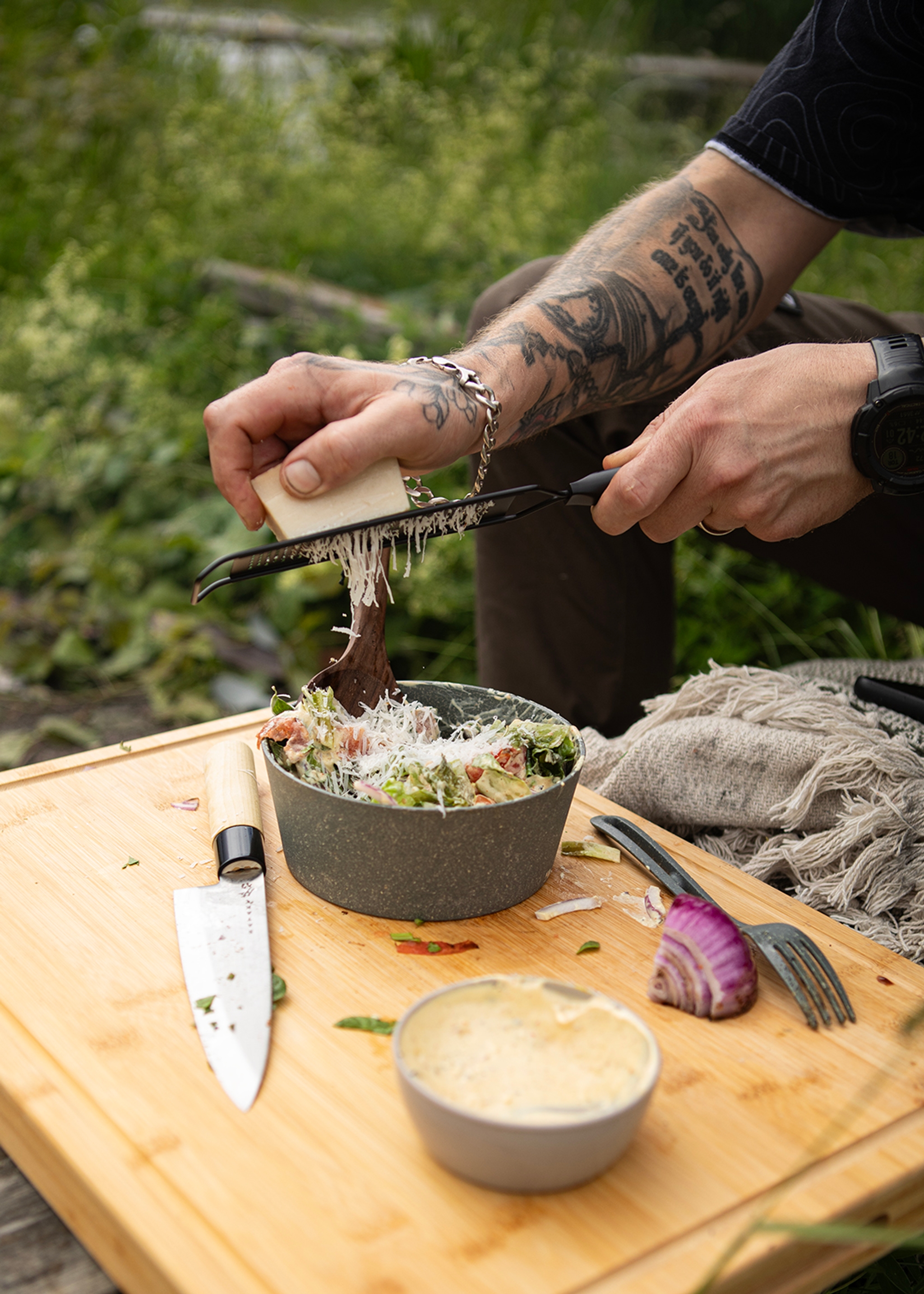 Tatuerad person river ost över en fräsch sallad utomhus på en träskärbräda.
