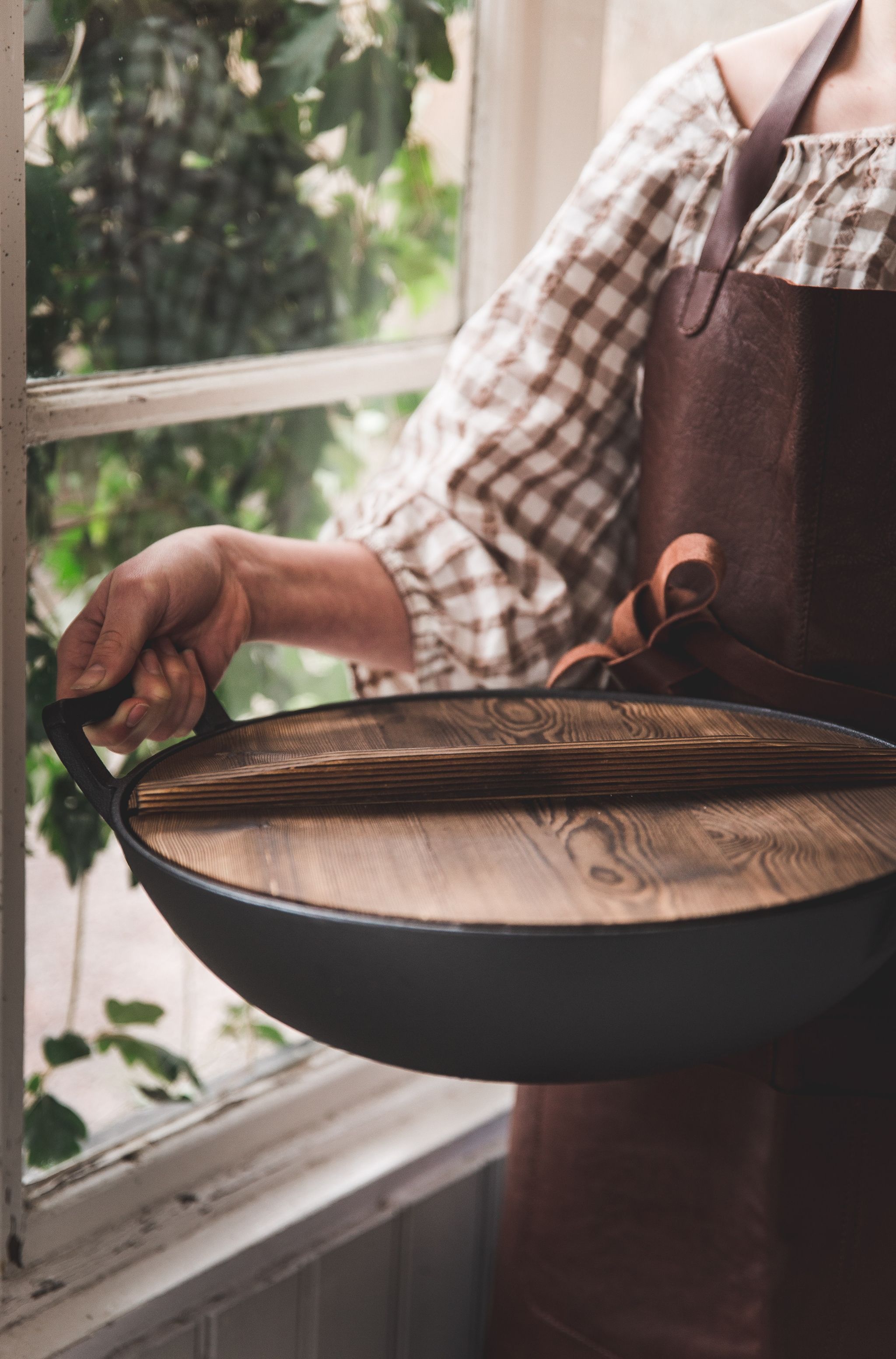 En person i ett brunt läderförkläde håller en svart panna med trälock, med ett fönster och gröna växter i bakgrunden.