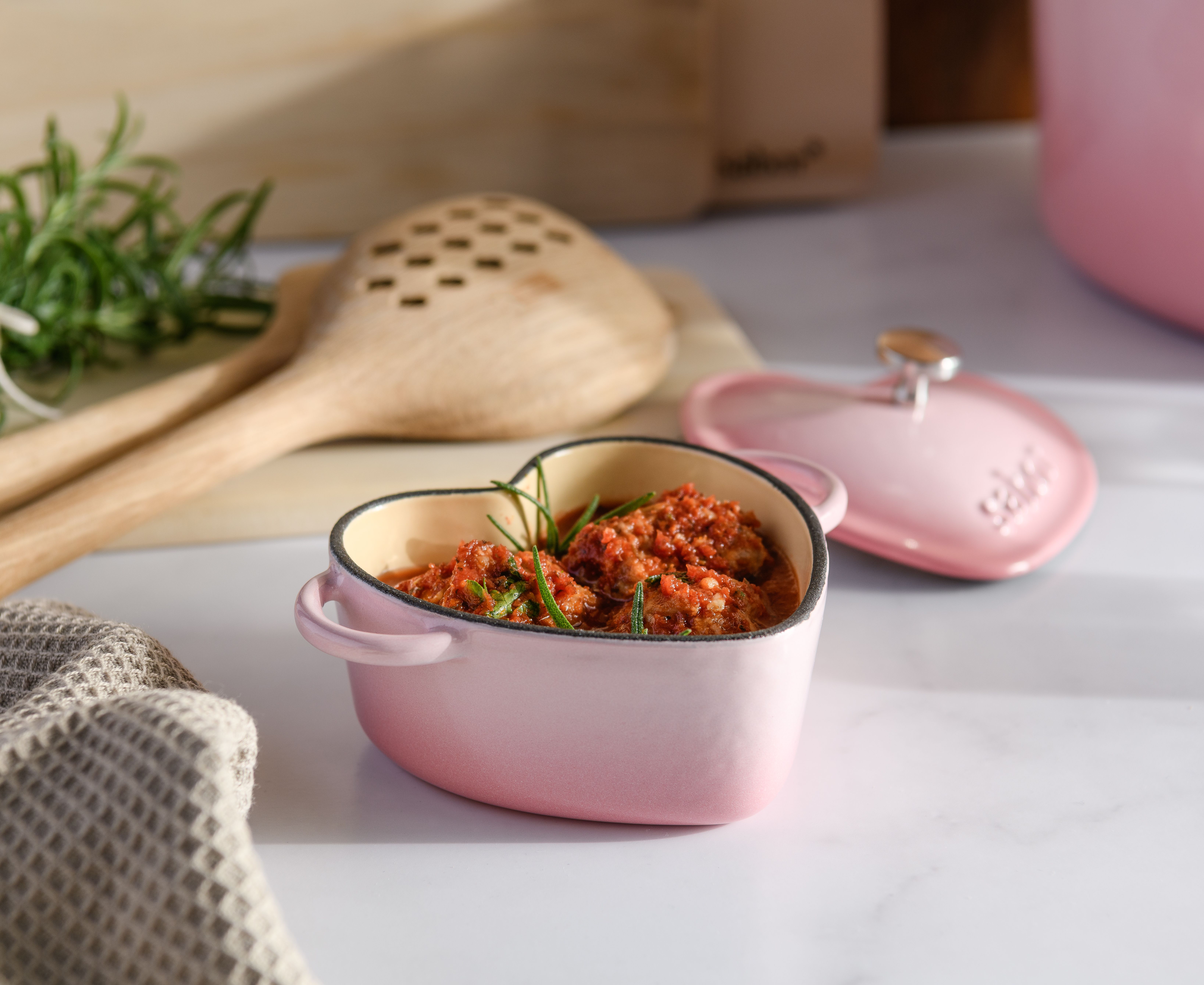 Rosa hjerteformet mini-gryte med rød stuing og rosmarin, ved siden av lokket og treskjeene.