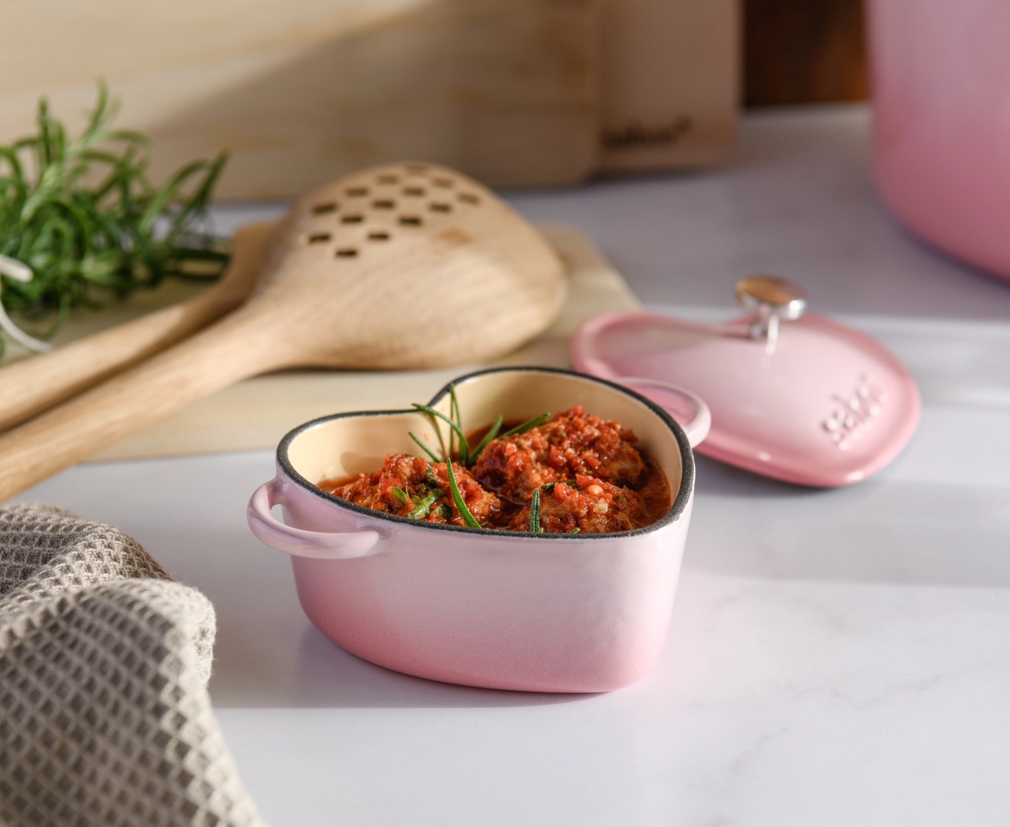 Rosa hjerteformet mini-gryte med rød stuing og rosmarin, ved siden av lokket og treskjeene.