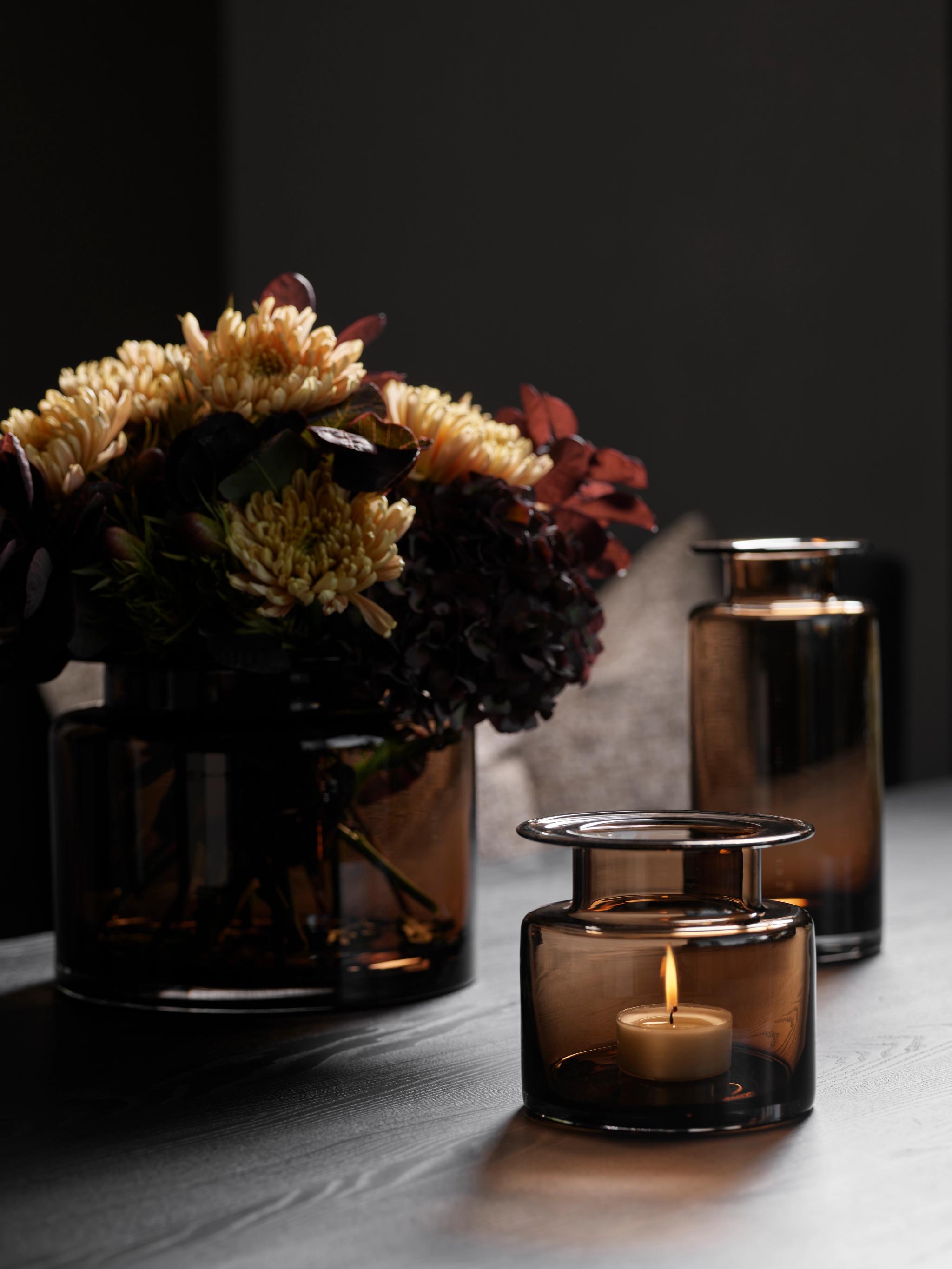 A dark brown glass vase of yellow and red flowers, a lit tea light in a matching glass holder, and another brown glass bottle on a dark table.