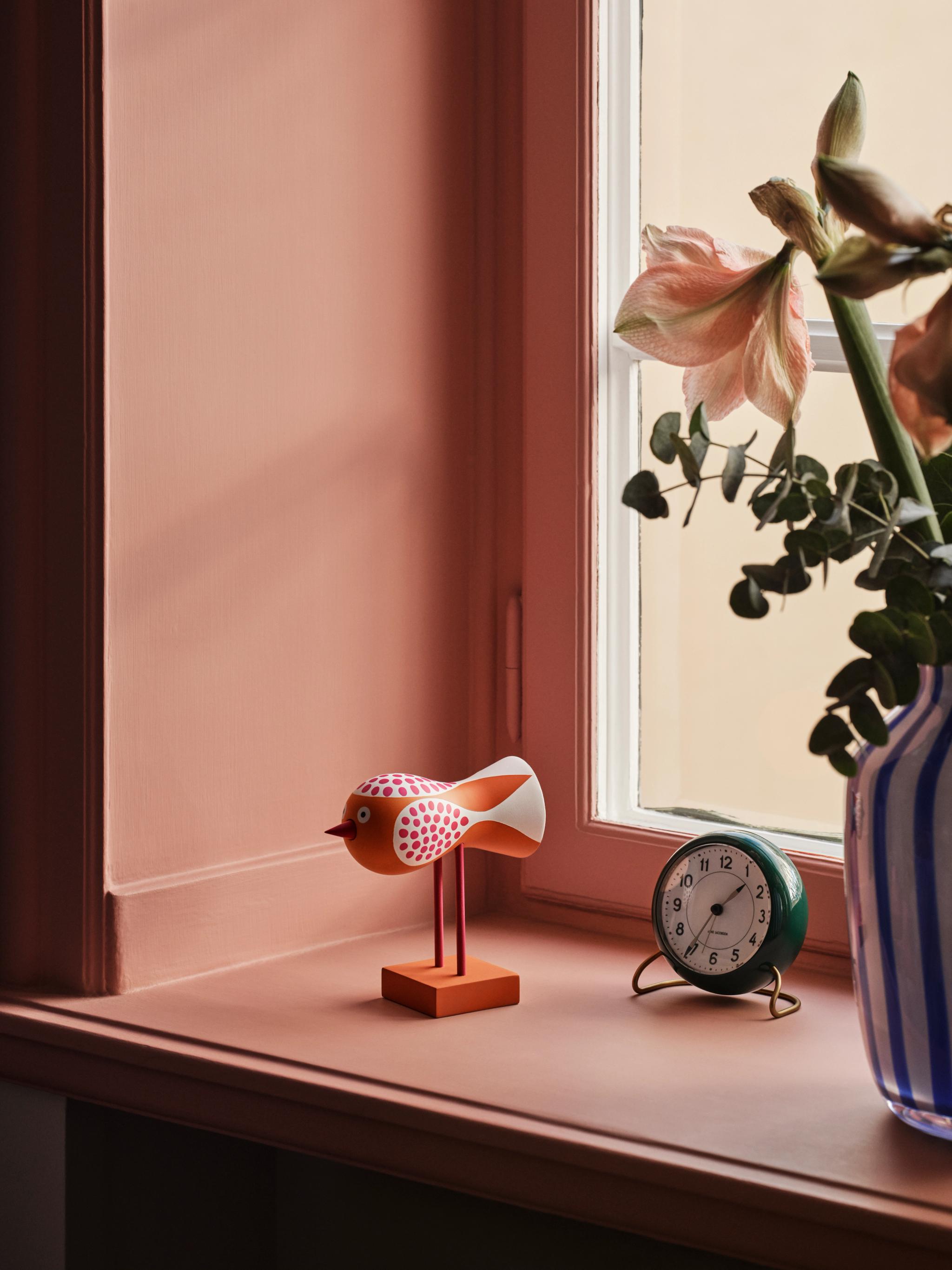 A terracotta-colored window sill holds an orange bird figurine, a green alarm clock, and a vase with flowers.