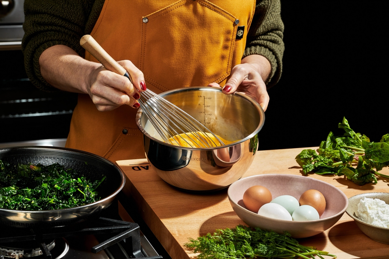 A person in an apron whisks eggs in a metal bowl, with a pan of cooked greens, fresh eggs, and herbs on a wooden counter.