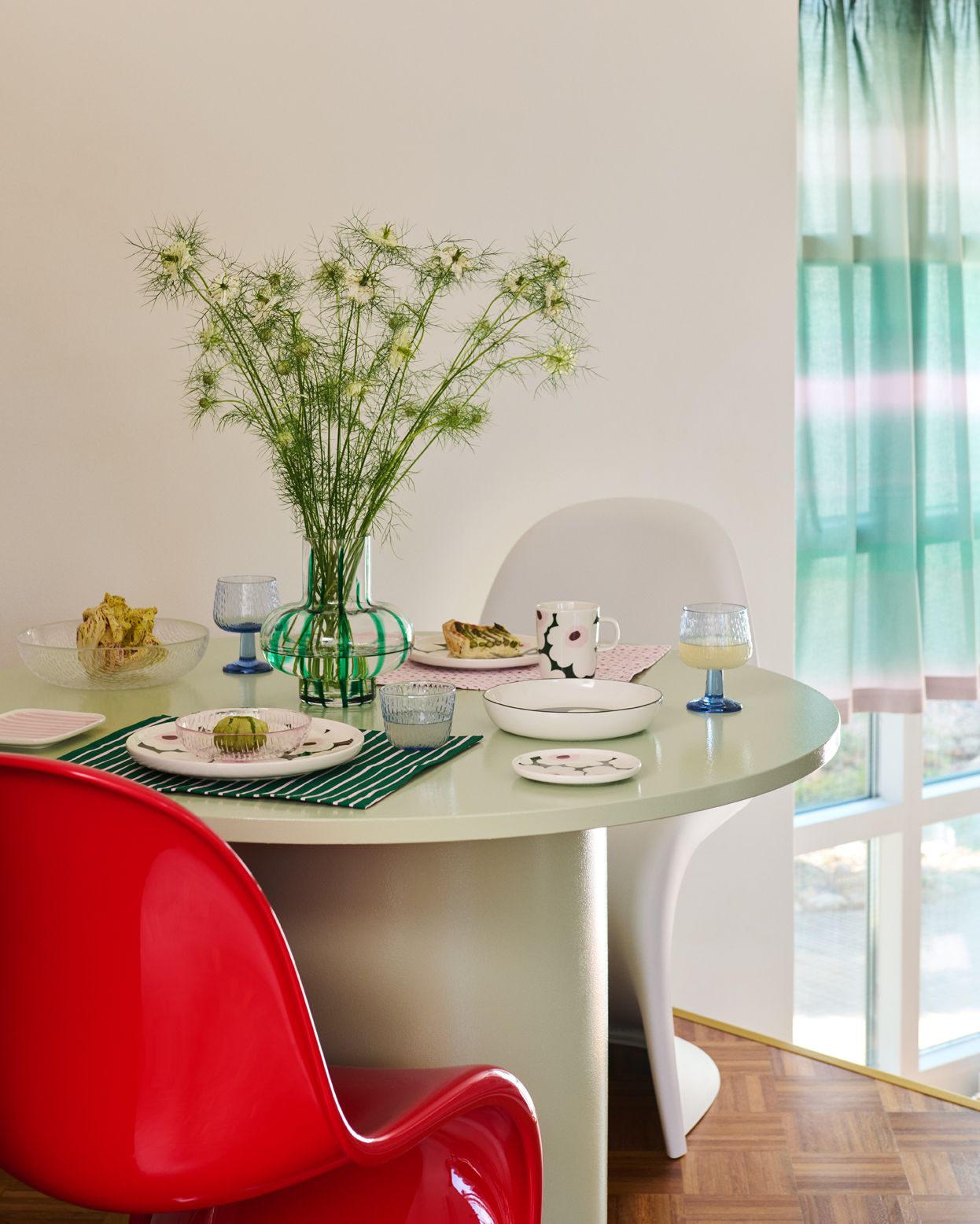 Ruokapöytä, jossa on vihreäraidallinen maljakko, jossa on vihreitä kasveja, quiche-viipale, kukkakuvioinen muki ja värikkäitä lasiesineitä.