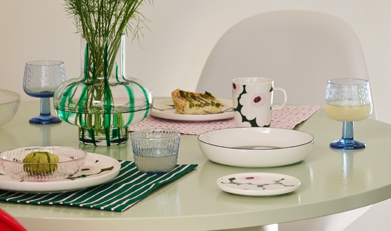 Vaaleanvihreä pyöreä ruokapöytä on katettu värikkäillä, kuviollisilla astioilla, kuten raidallinen maljakko vihreillä kasveilla, kukkamuki ja siivu quichea, etualalla punaisen tuolin kanssa.