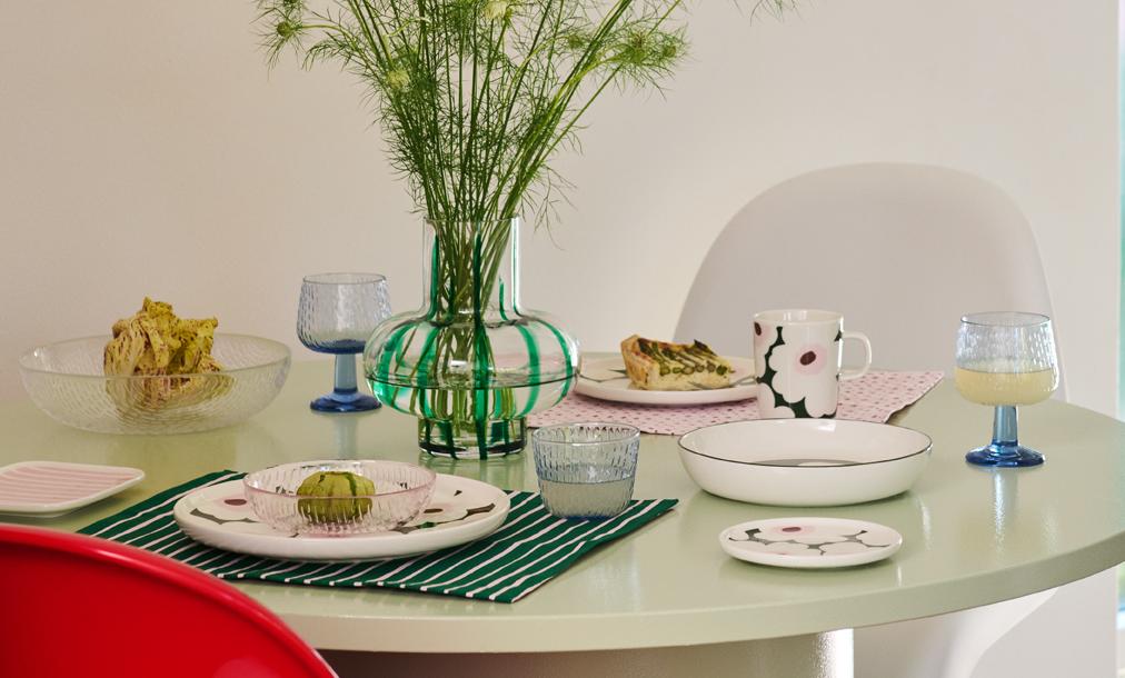 Vaaleanvihreä pyöreä ruokapöytä on katettu värikkäillä, kuviollisilla astioilla, kuten raidallinen maljakko vihreillä kasveilla, kukkamuki ja siivu quichea, etualalla punaisen tuolin kanssa.