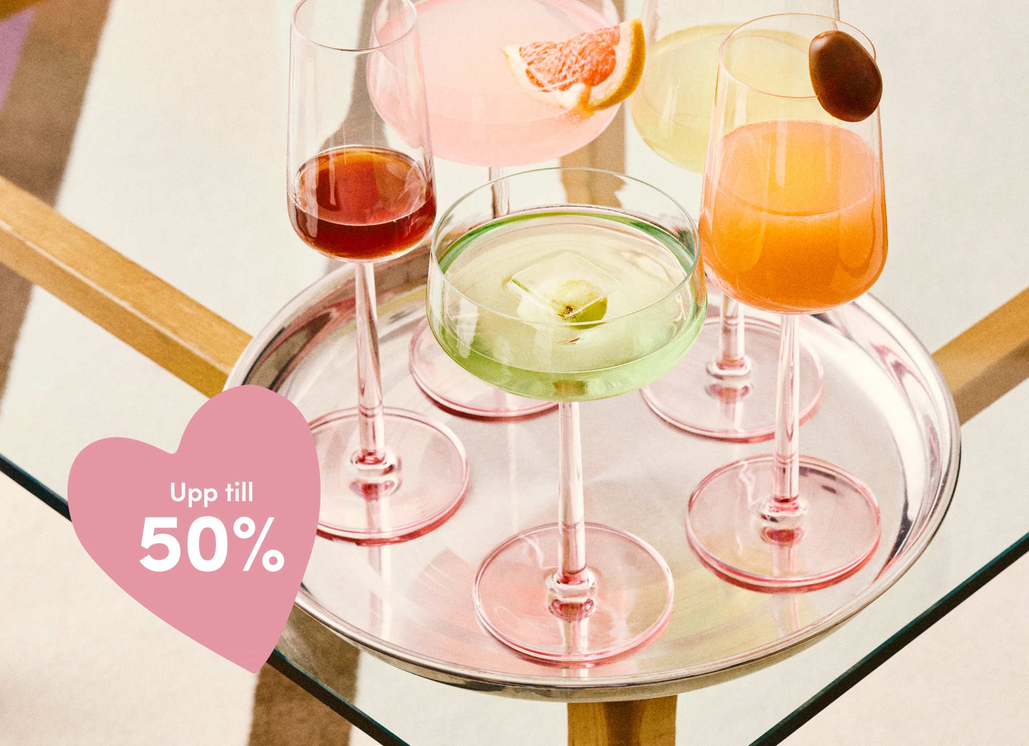 Blandade cocktails i glas med rosa fot på en bricka med en hjärtformad text "Upp till 50 %" rabatt.