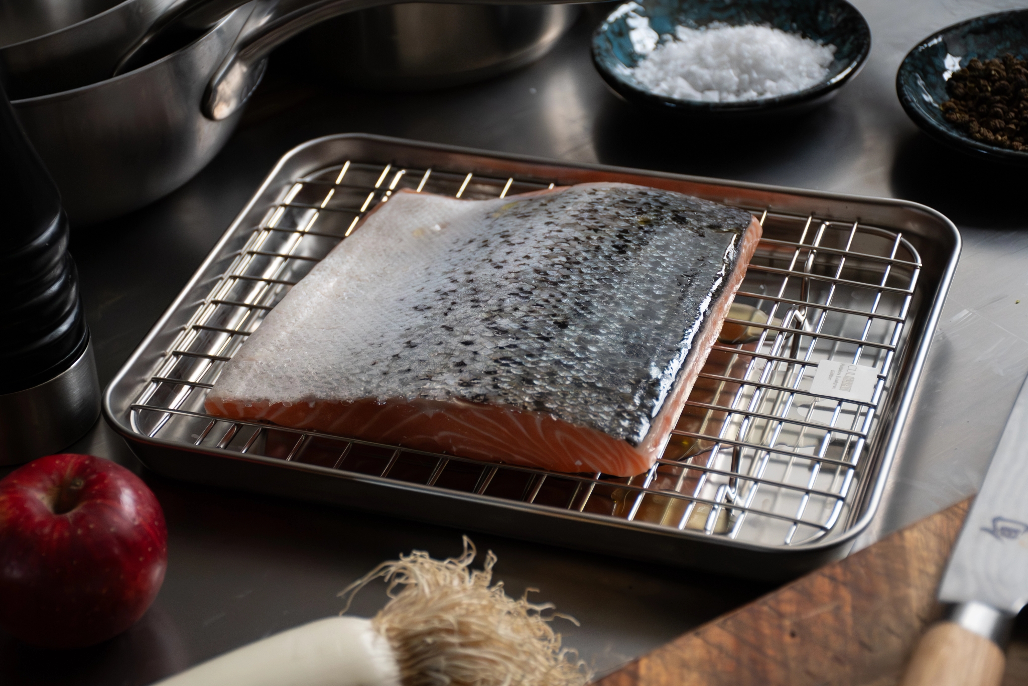 Rå laxfilé på ett metallgaller över en vätskefylld bricka, omgiven av salt, peppar, ett äpple och en purjolök på en bänk.