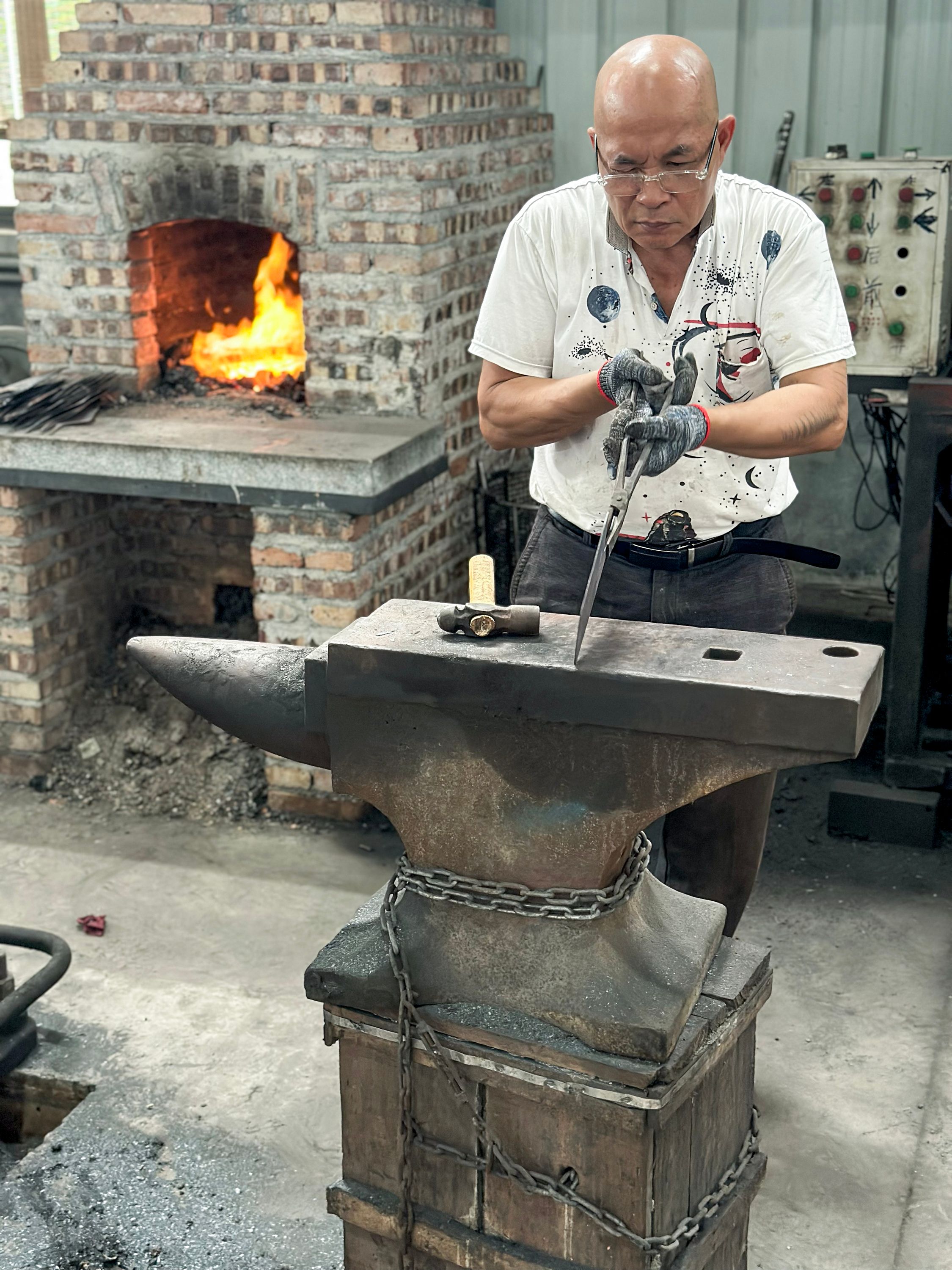 En smed bearbetar ett metallstycke på ett städ med en glödande smedja i bakgrunden.