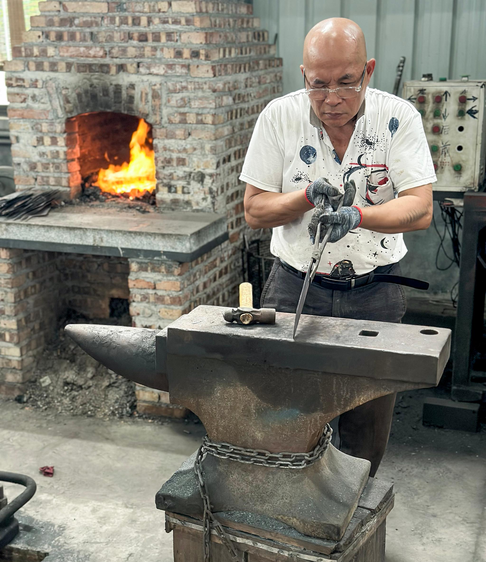 En smed bearbetar ett metallstycke på ett städ med en glödande smedja i bakgrunden.