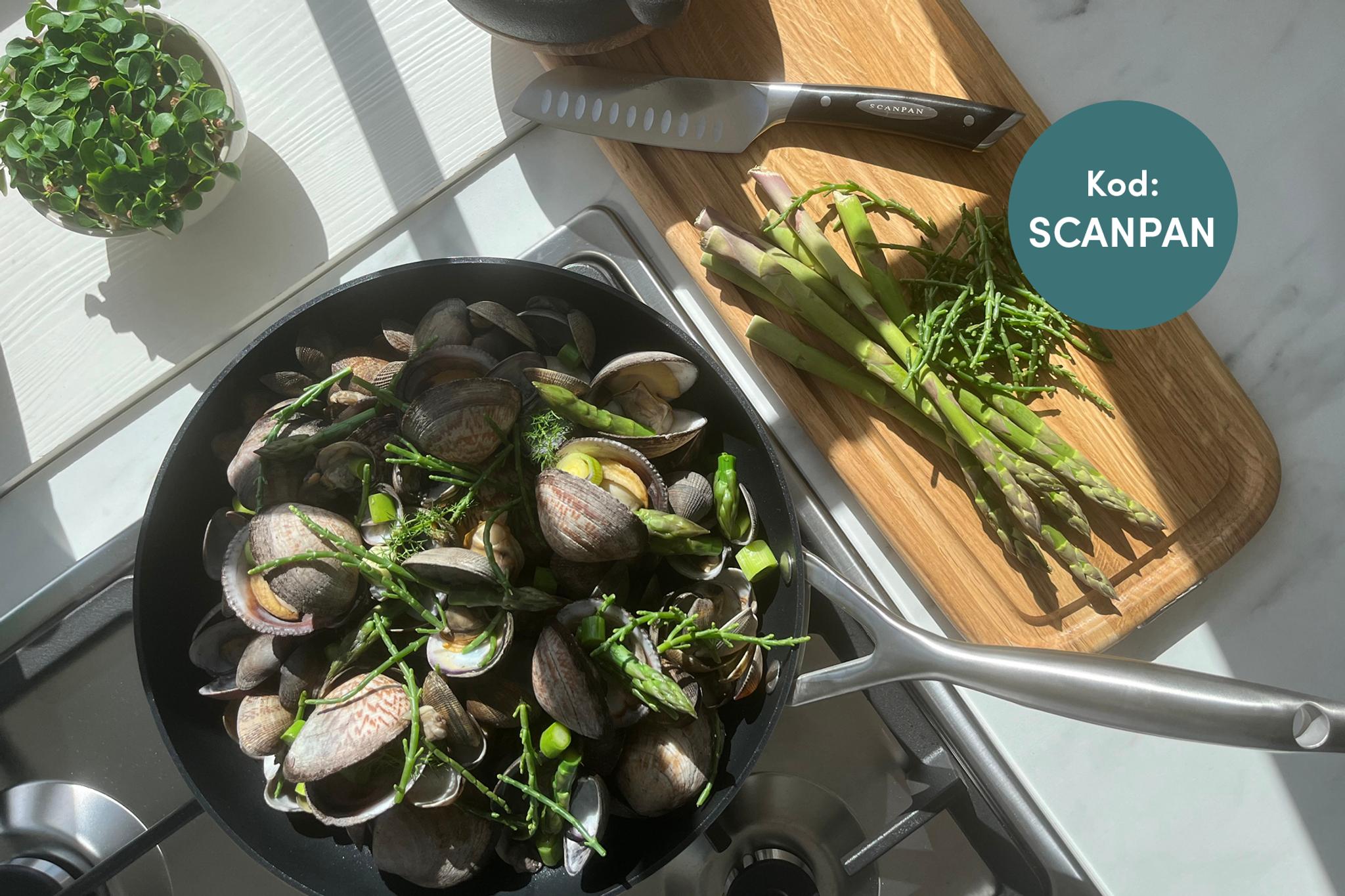 A pan of cooked clams, asparagus, and greens on a stovetop next to a cutting board with raw asparagus and a knife. A 'SCANPAN' logo is visible.