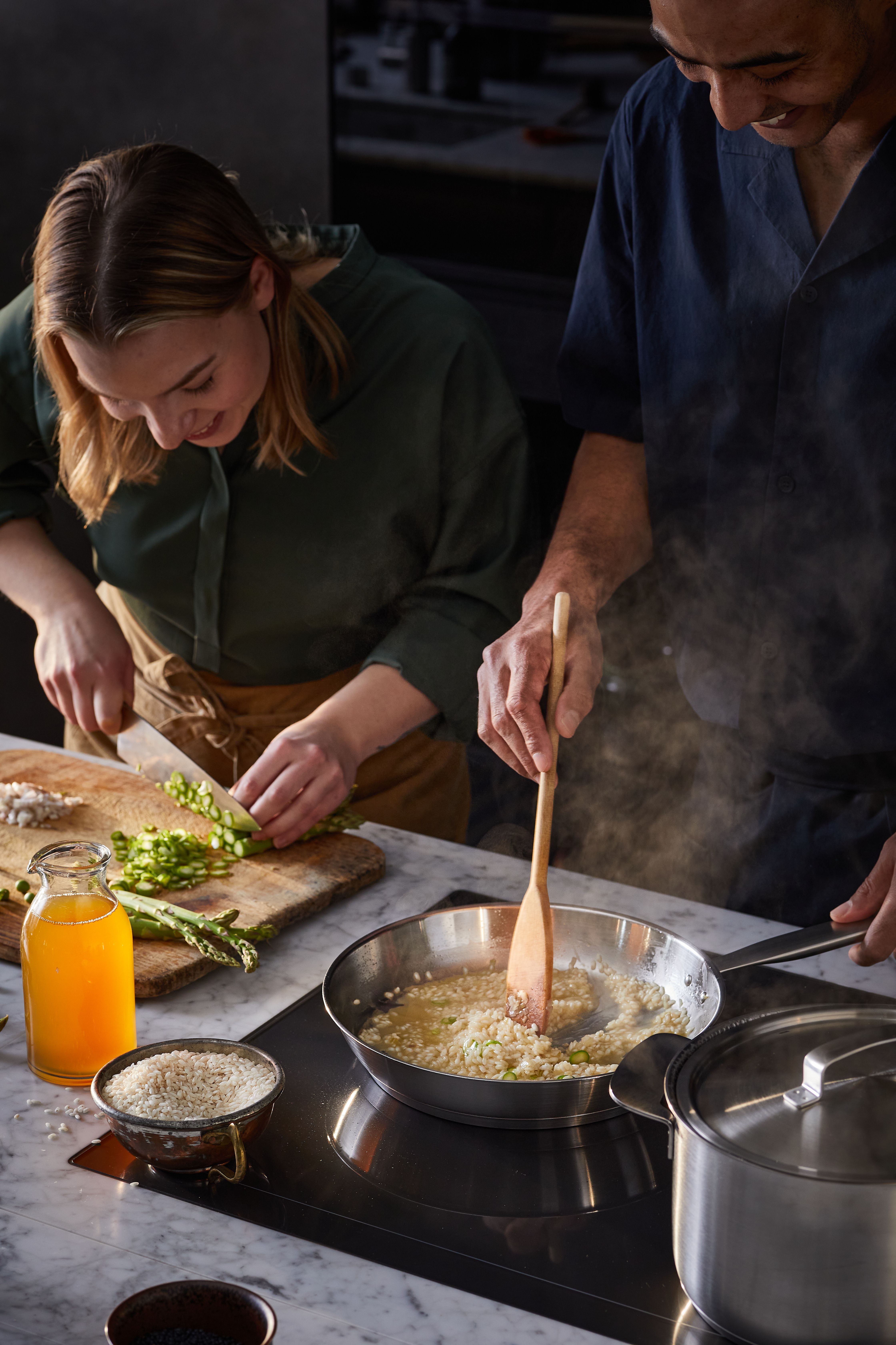 Nainen pilkkoo parsaa, kun taas mies sekoittaa risottoa höyryävällä pannulla induktioliesitasolla.