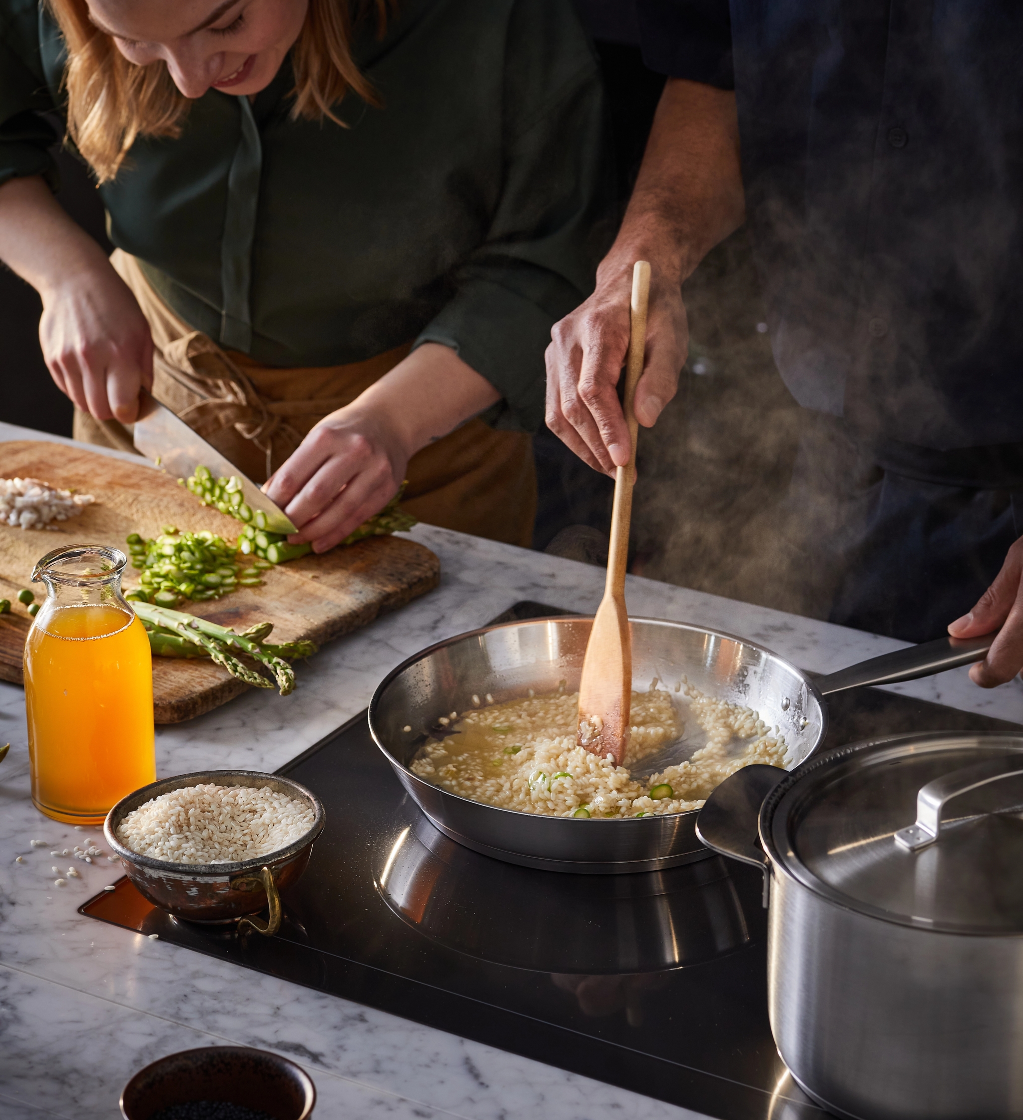 Nainen pilkkoo parsaa, kun taas mies sekoittaa risottoa höyryävällä pannulla induktioliesitasolla.