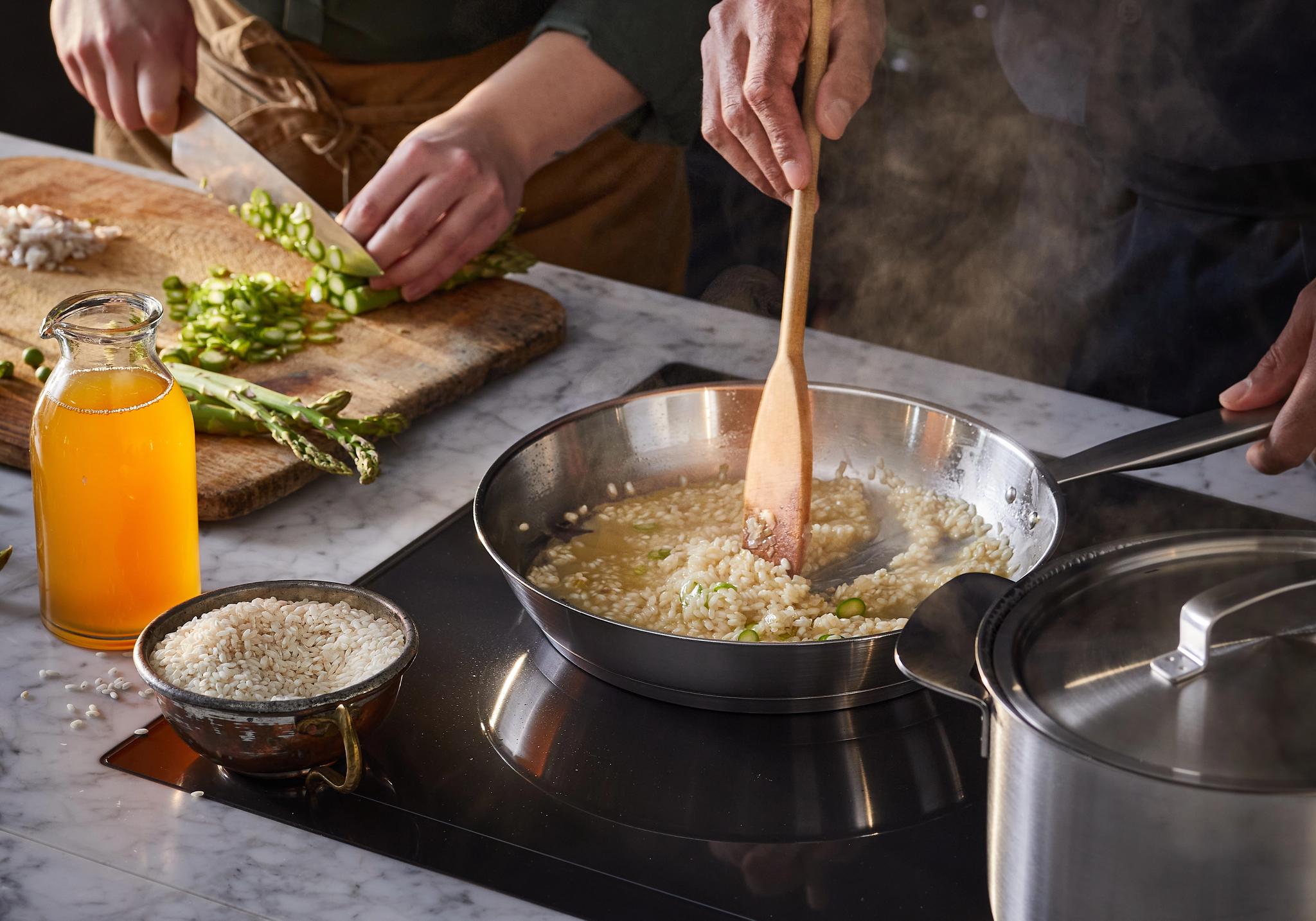 Kaksi ihmistä kokkaa: toinen pilkkoo parsaa, toinen sekoittaa risottoa höyryävällä pannulla.