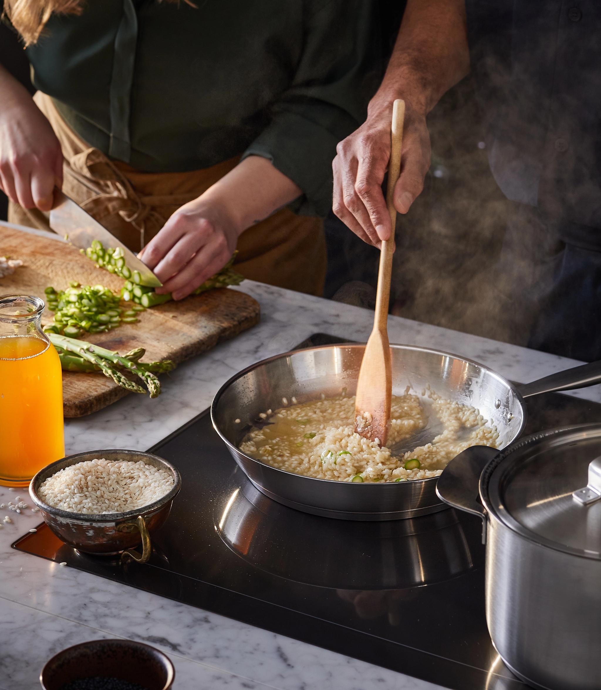 Kaksi ihmistä kokkaa: toinen pilkkoo parsaa, toinen sekoittaa risottoa höyryävällä pannulla.