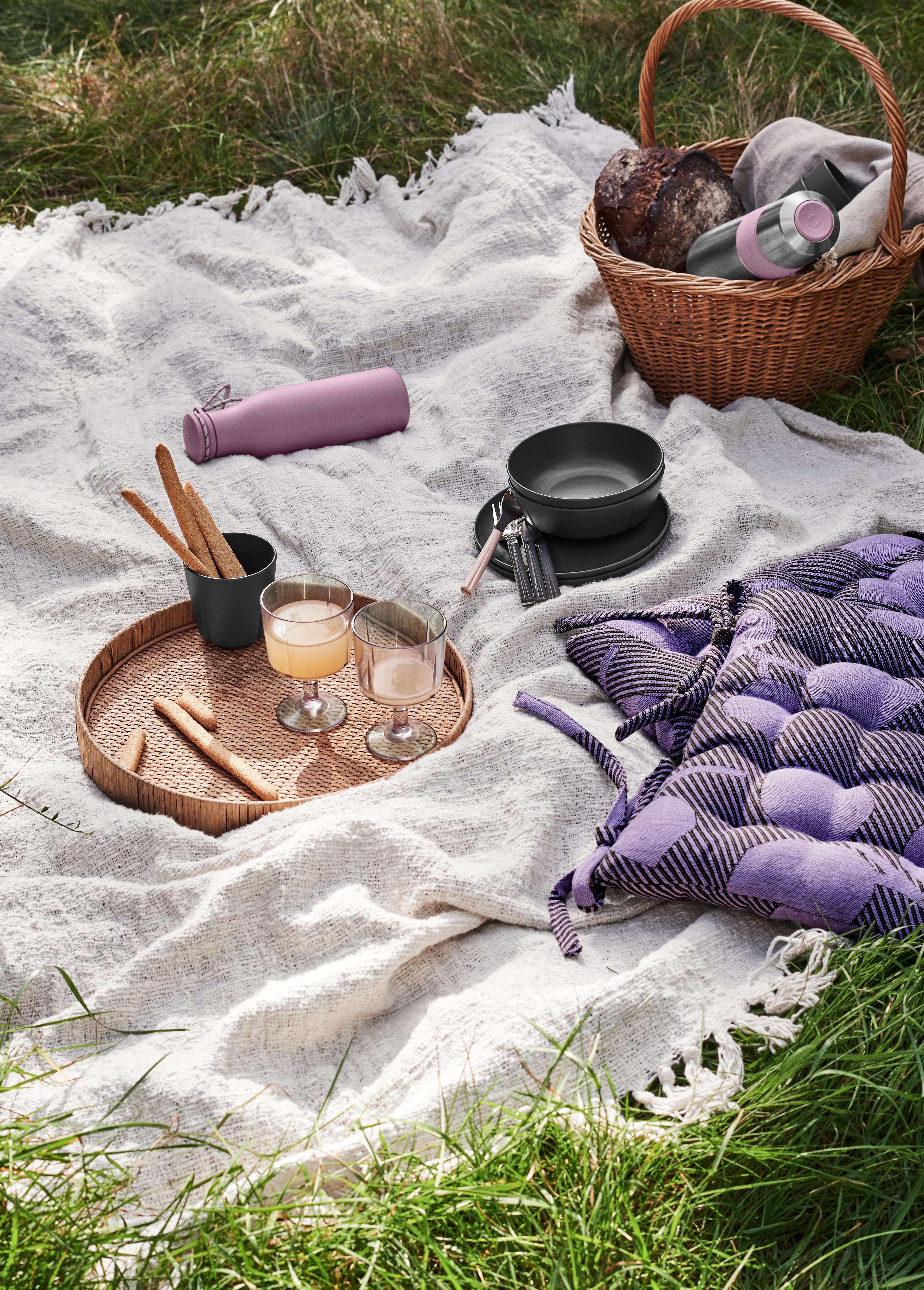 Somrig picknick på filt i gräset med korg, dryck, bröd och färgglada kuddar.