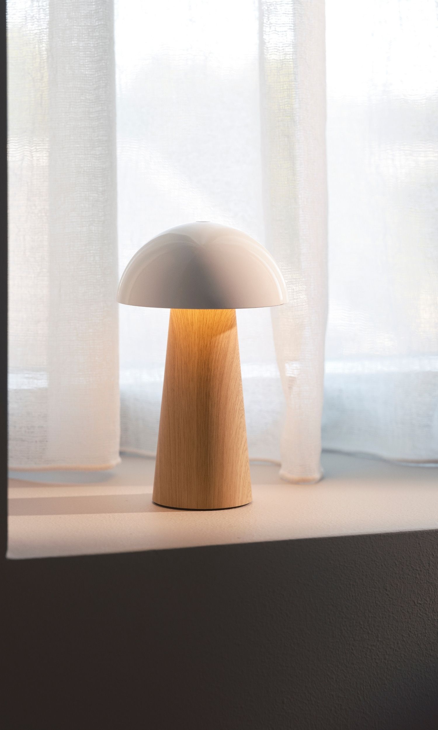 A modern mushroom-shaped lamp with a white shade and light wood base glows on a windowsill in front of sheer white curtains.