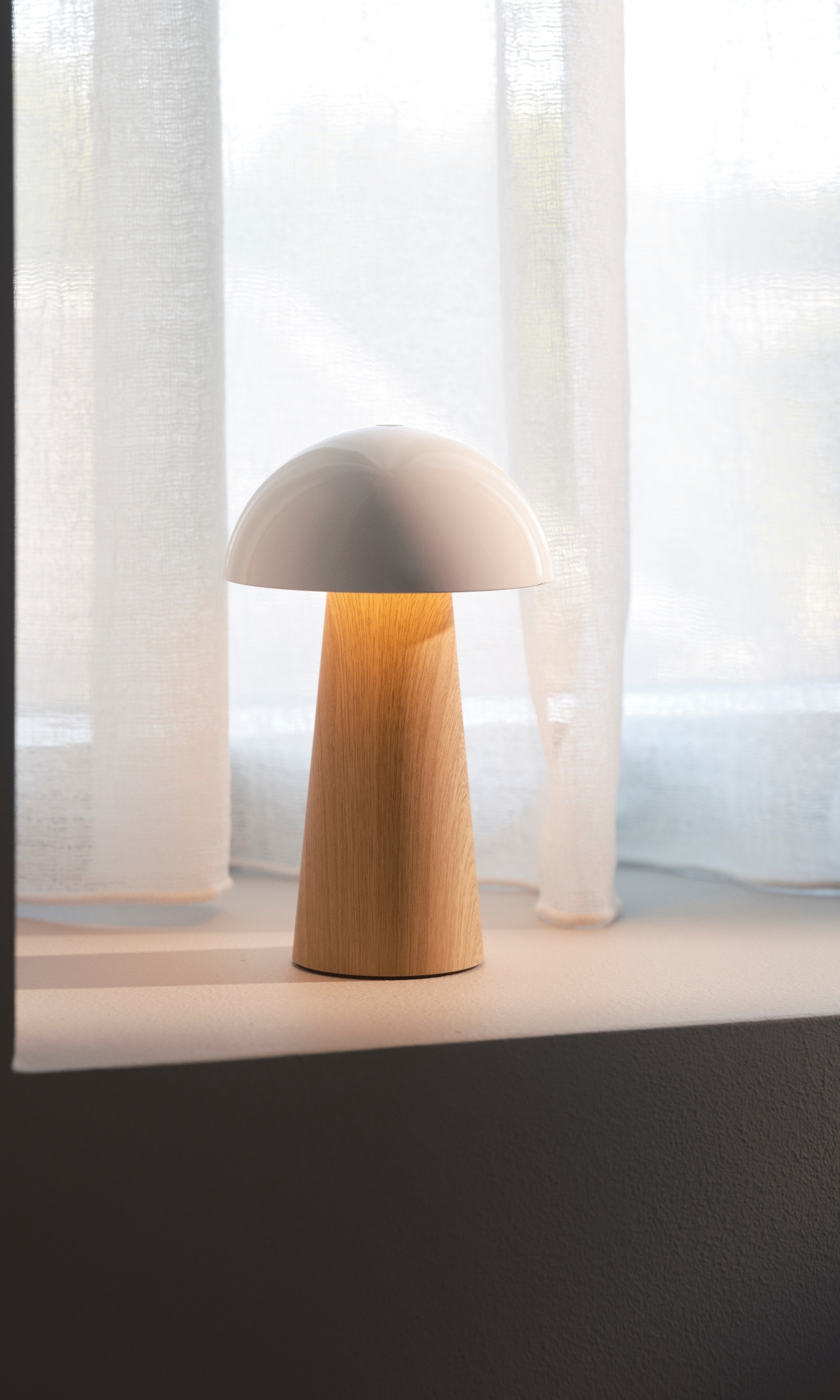 A modern mushroom-shaped lamp with a white cap and wooden base sits on a shelf in front of sheer white curtains.