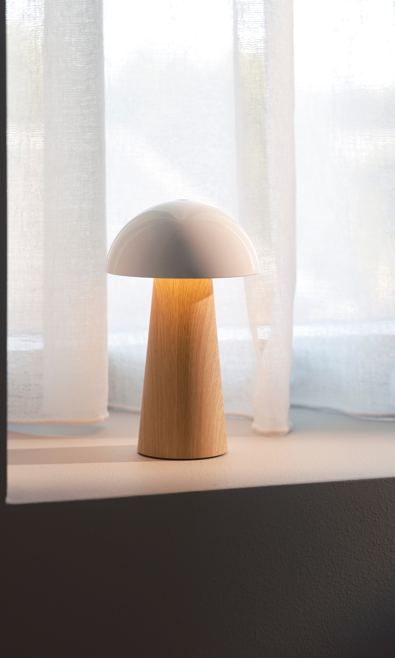 A modern mushroom-shaped lamp with a white cap and wooden base sits on a shelf in front of sheer white curtains.