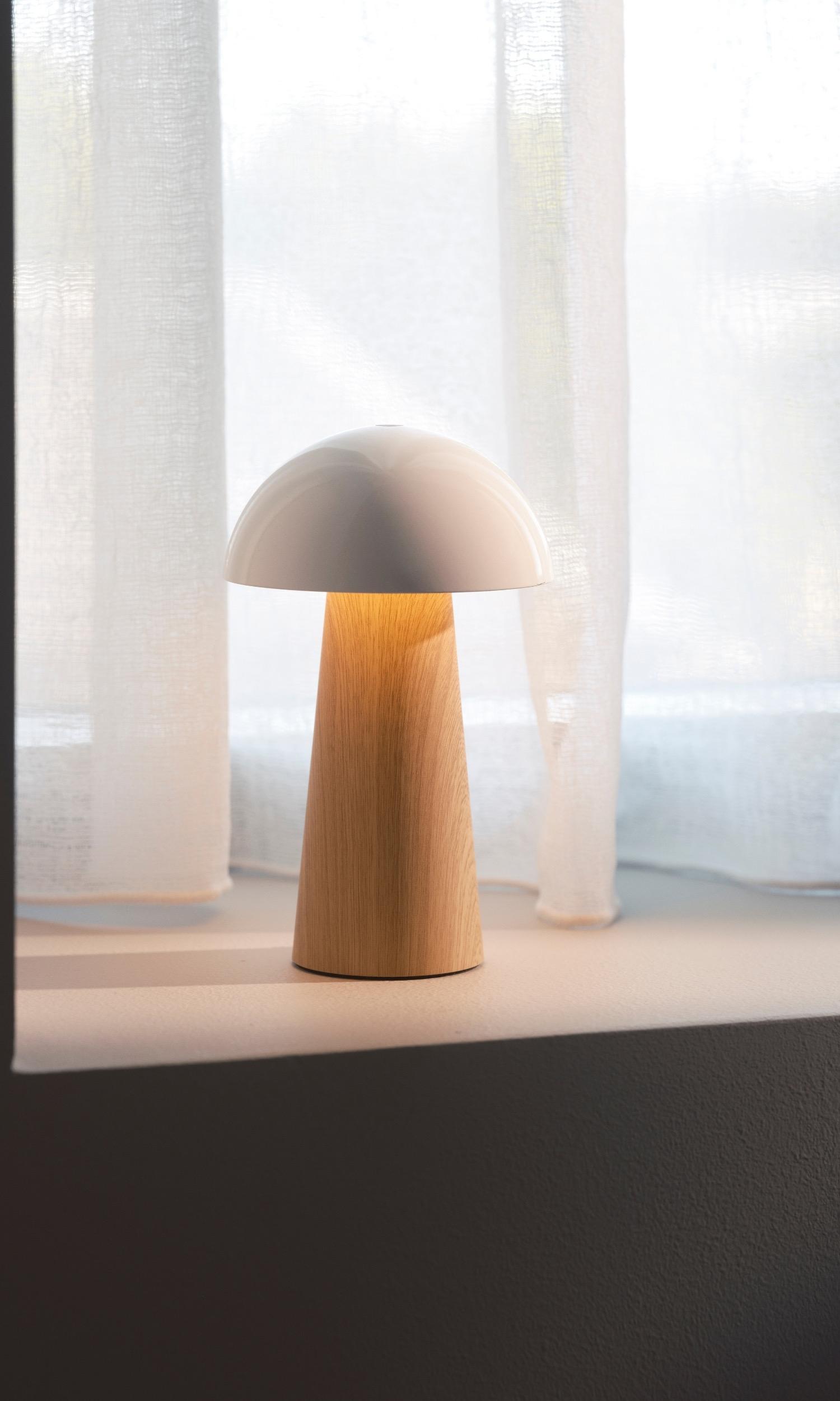 A modern mushroom-shaped lamp with a light wood base and white shade sits on a windowsill with sheer white curtains behind it.