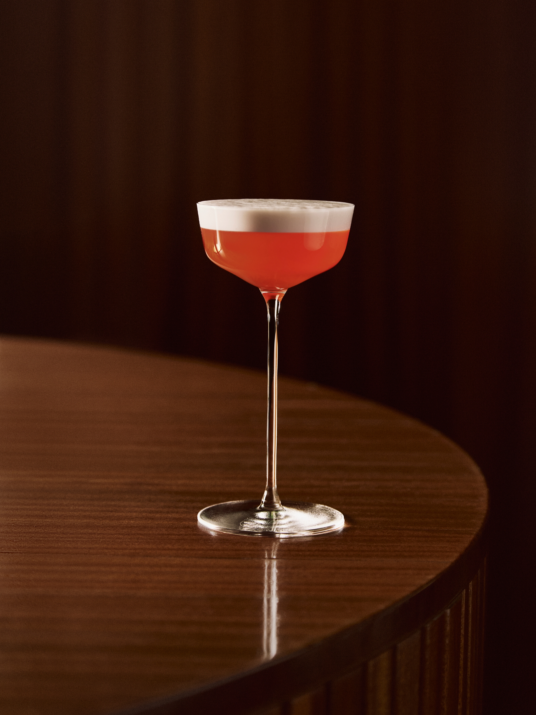 En röd cocktail med vitt skum i ett coupéglas på ett träbord.