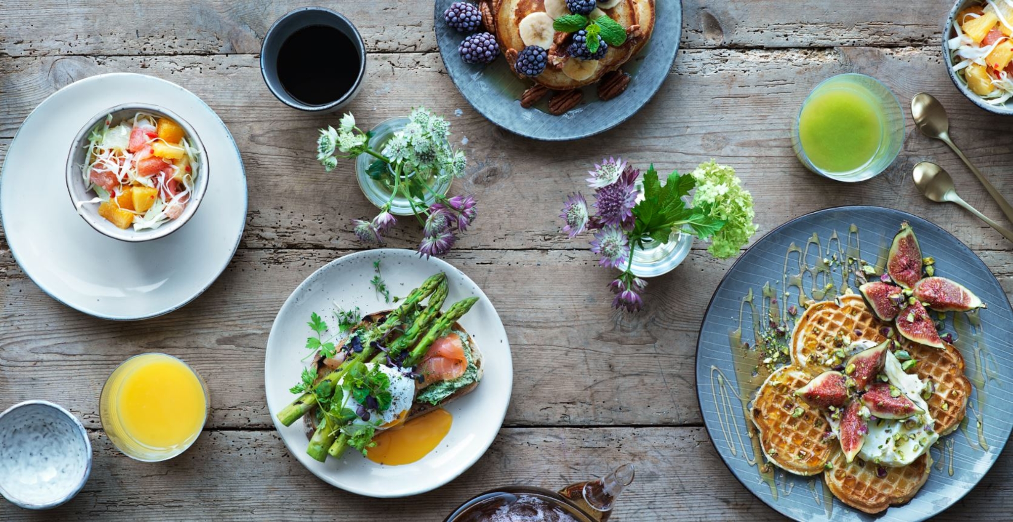Färgglad brunch med våfflor, färska fikon, sparris, lax, frukt, kaffe och juice på ett rustikt träbord.