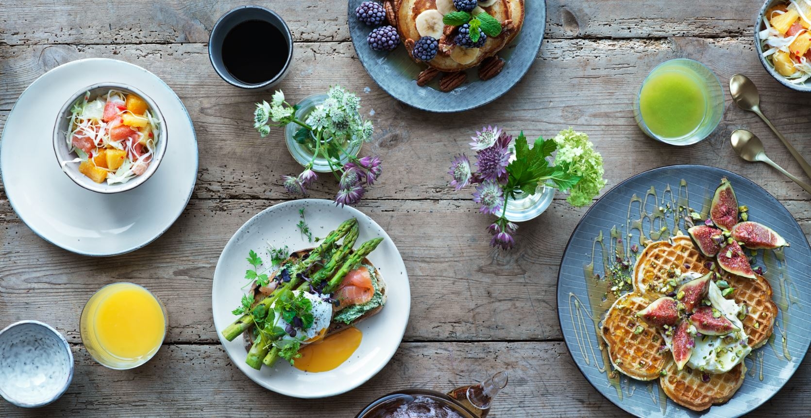 Färgglad brunch med våfflor, färska fikon, sparris, lax, frukt, kaffe och juice på ett rustikt träbord.