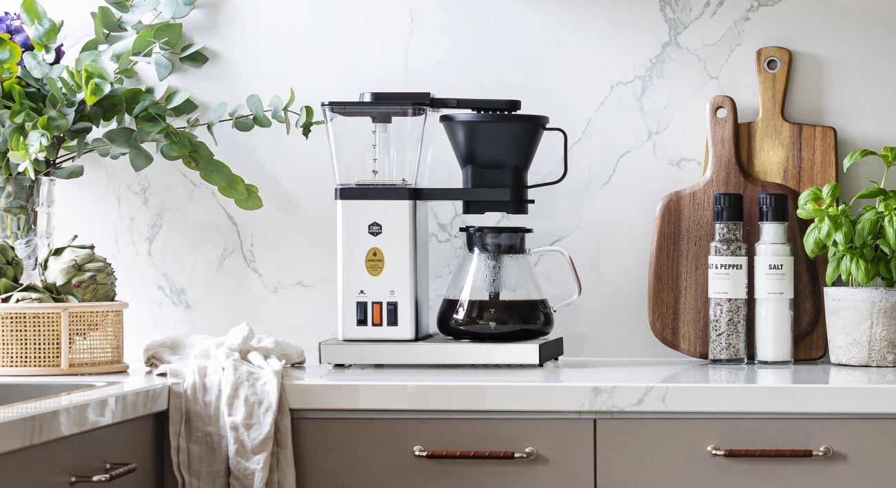 En kaffetrakter på en lys kjøkkenbenk med marmorbakplate, planter og skjærefjøler i tre.