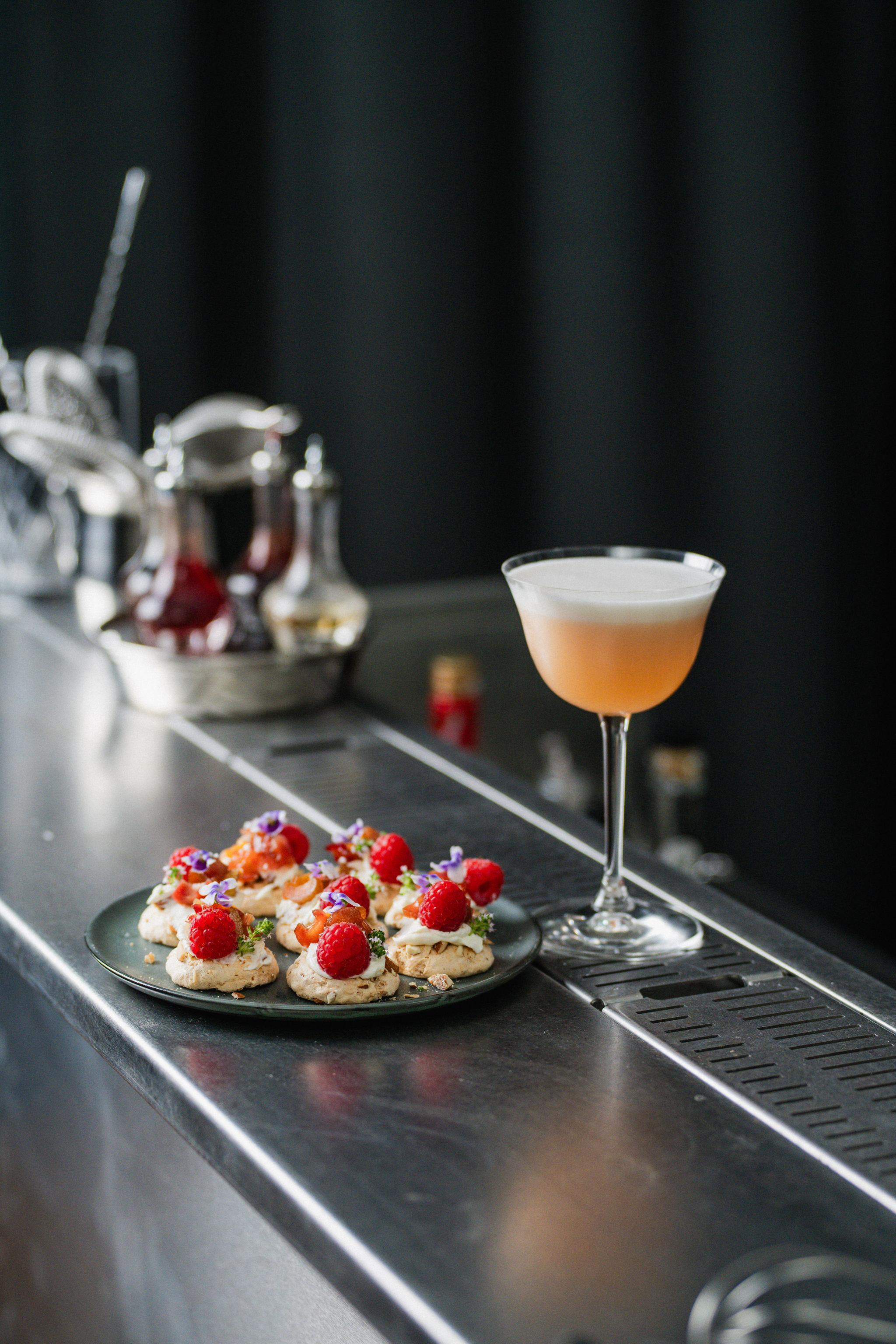 Elegant desserttallrik med små bakverk toppade med hallon och ätbara blommor bredvid en skummig cocktail på en bardisk.