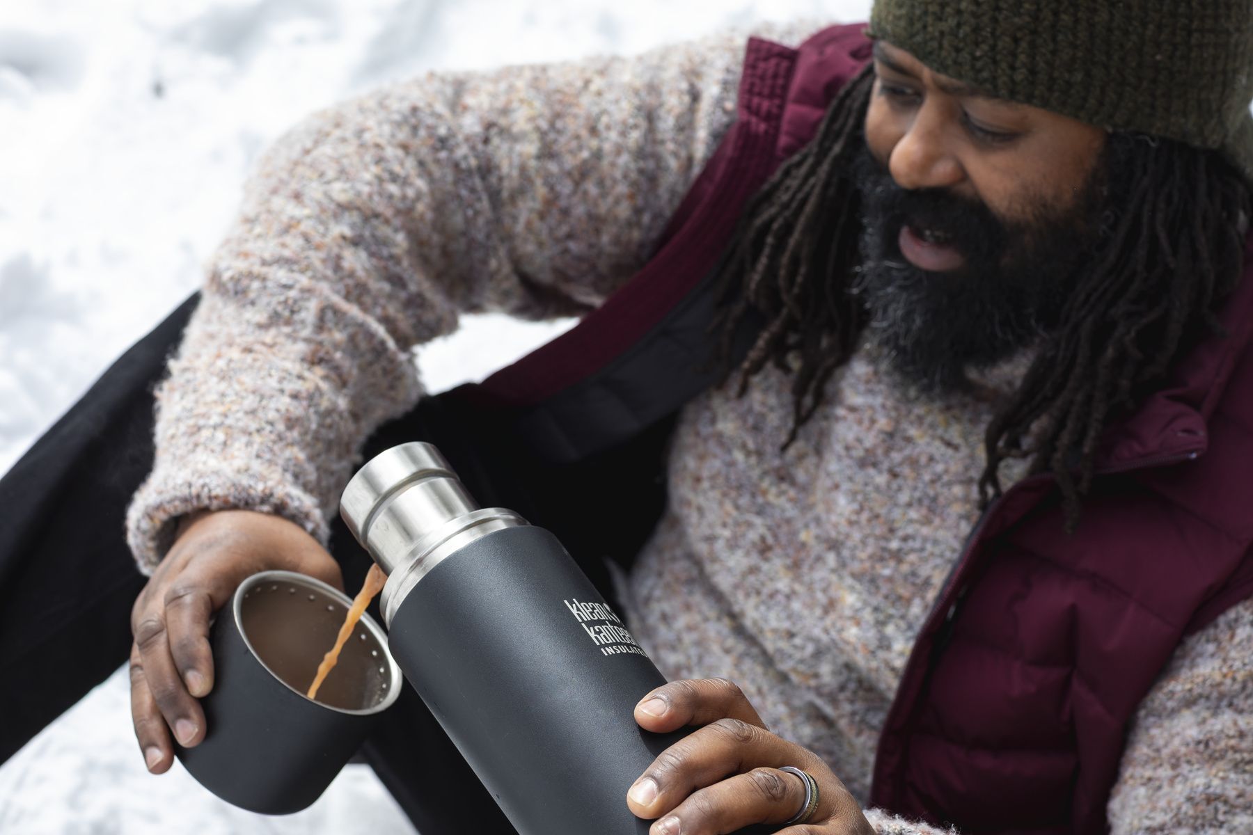 En person med dreadlocks och skägg häller varm choklad från en svart termos i en kopp i snön.