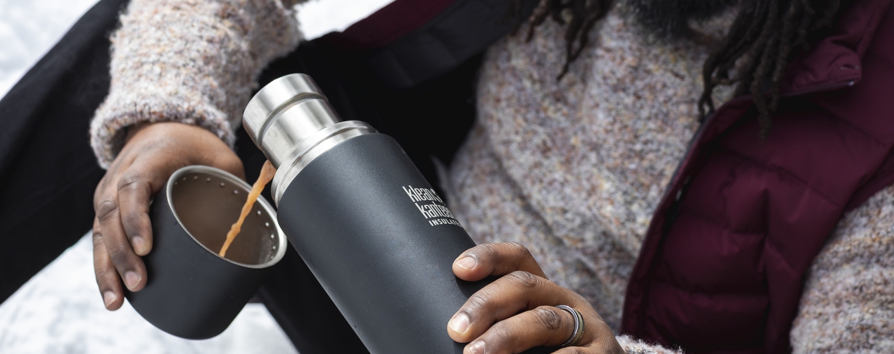 En person i mössa och väst häller en varm dryck från en svart termos i dess kopp i snön.