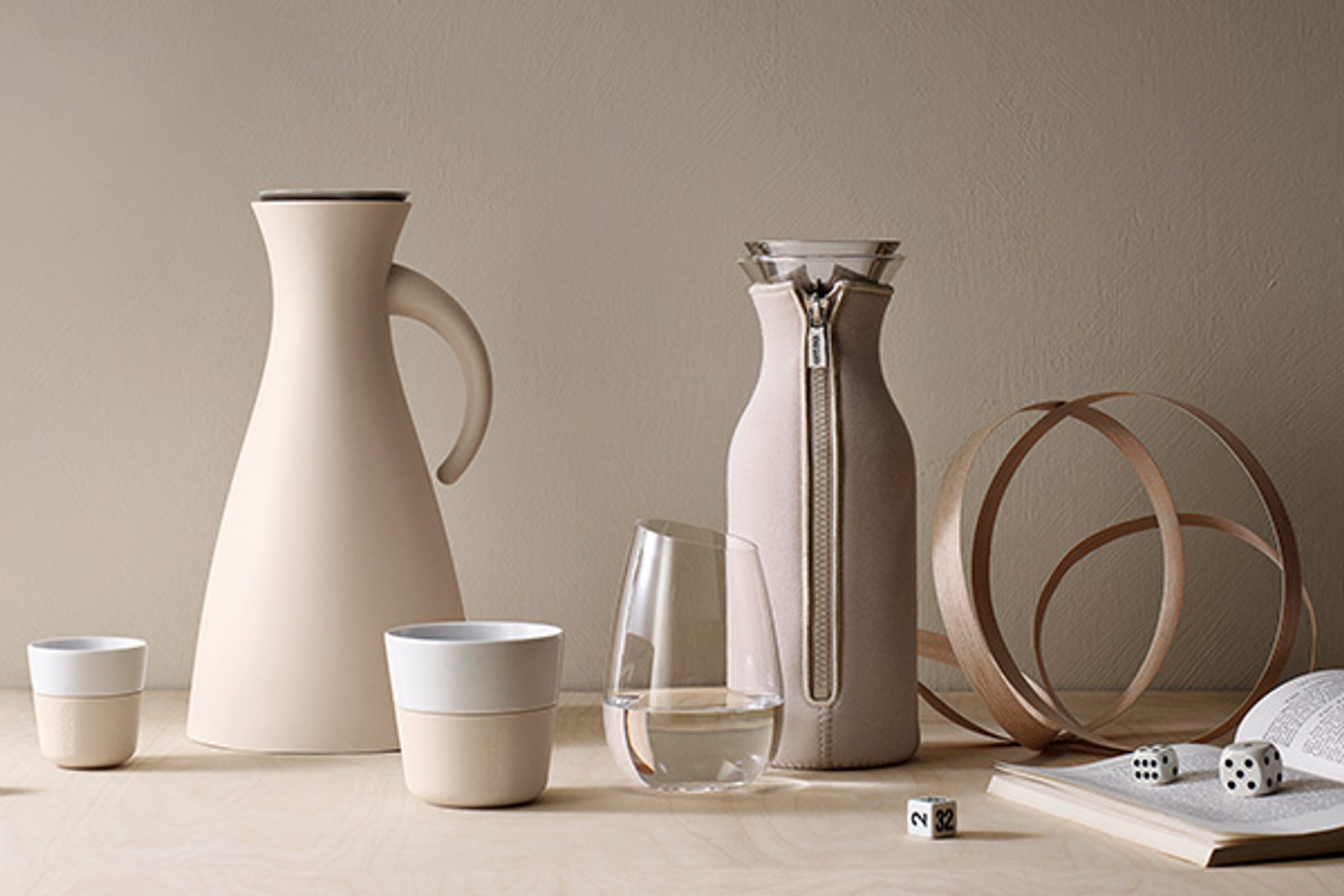 A minimalist still life featuring beige home decor: a thermal carafe, two ceramic cups, a water glass, a sleeved carafe, a wooden sculpture, an open book, and dice on a light wood table.