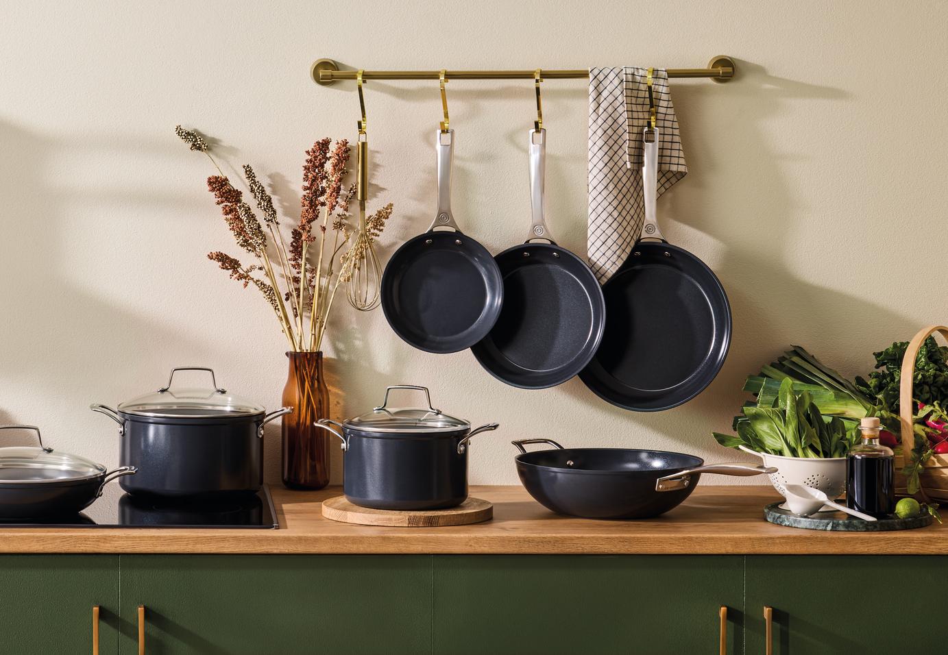 Mörkgrå grytor och stekpannor hänger på en köksbänk, omgivna av färska råvaror och inredning.