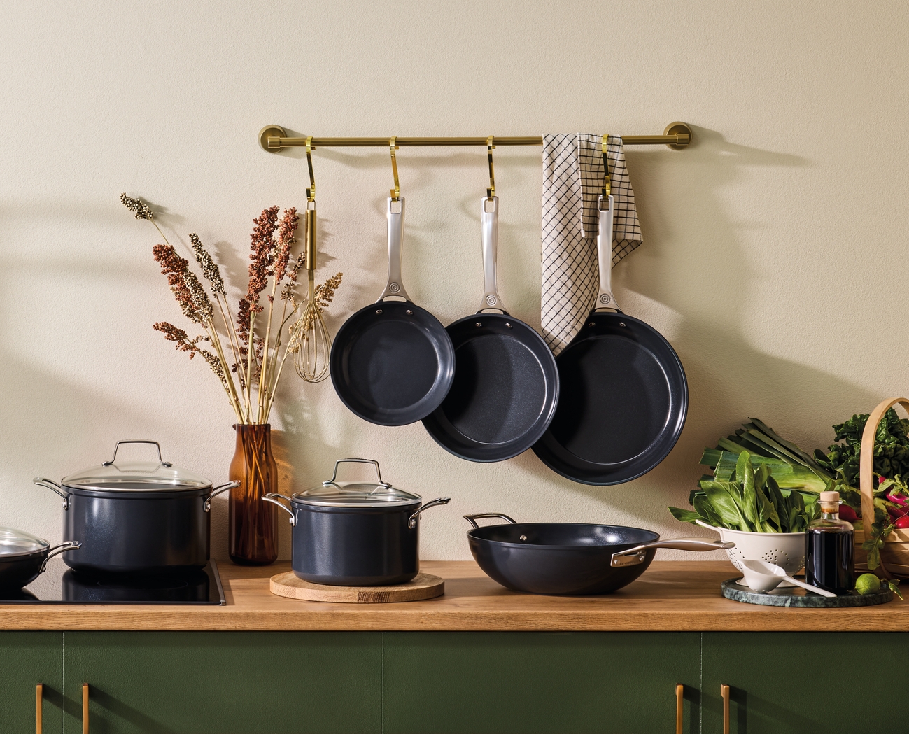 Mörkgrå grytor och stekpannor hänger på en köksbänk, omgivna av färska råvaror och inredning.