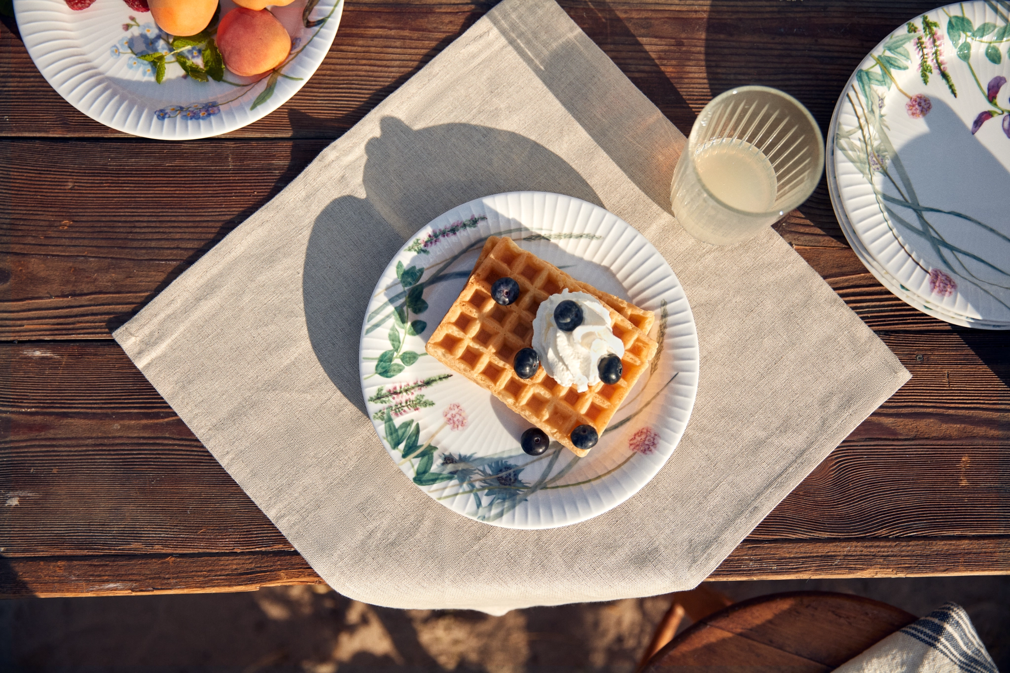 Vohveli kermavaahdolla ja mustikoilla kukkaisella lautasella, tarjoiltuna puupöydällä ulkona aprikoosien ja juomien kanssa.