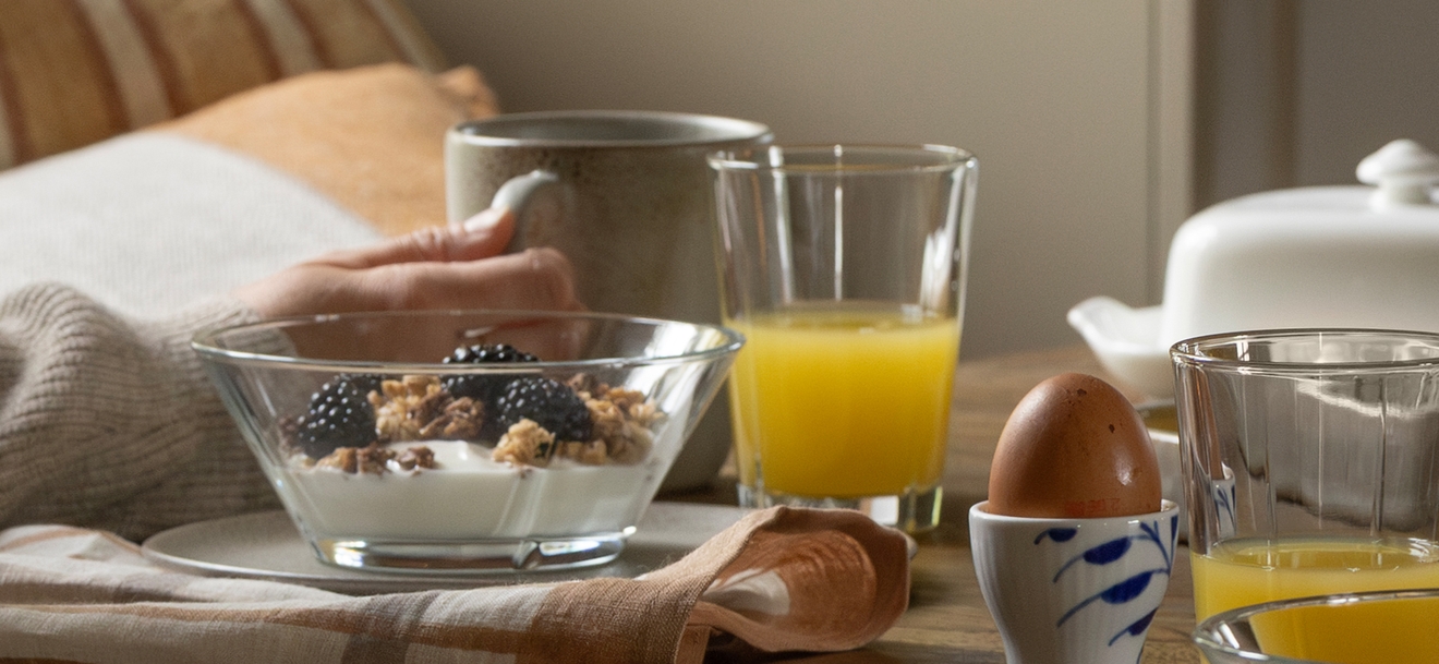 Kvinna i mysig tröja sitter vid ett fönster och äter frukost med kaffe, juice och müsli i ett varmt inrett kök.