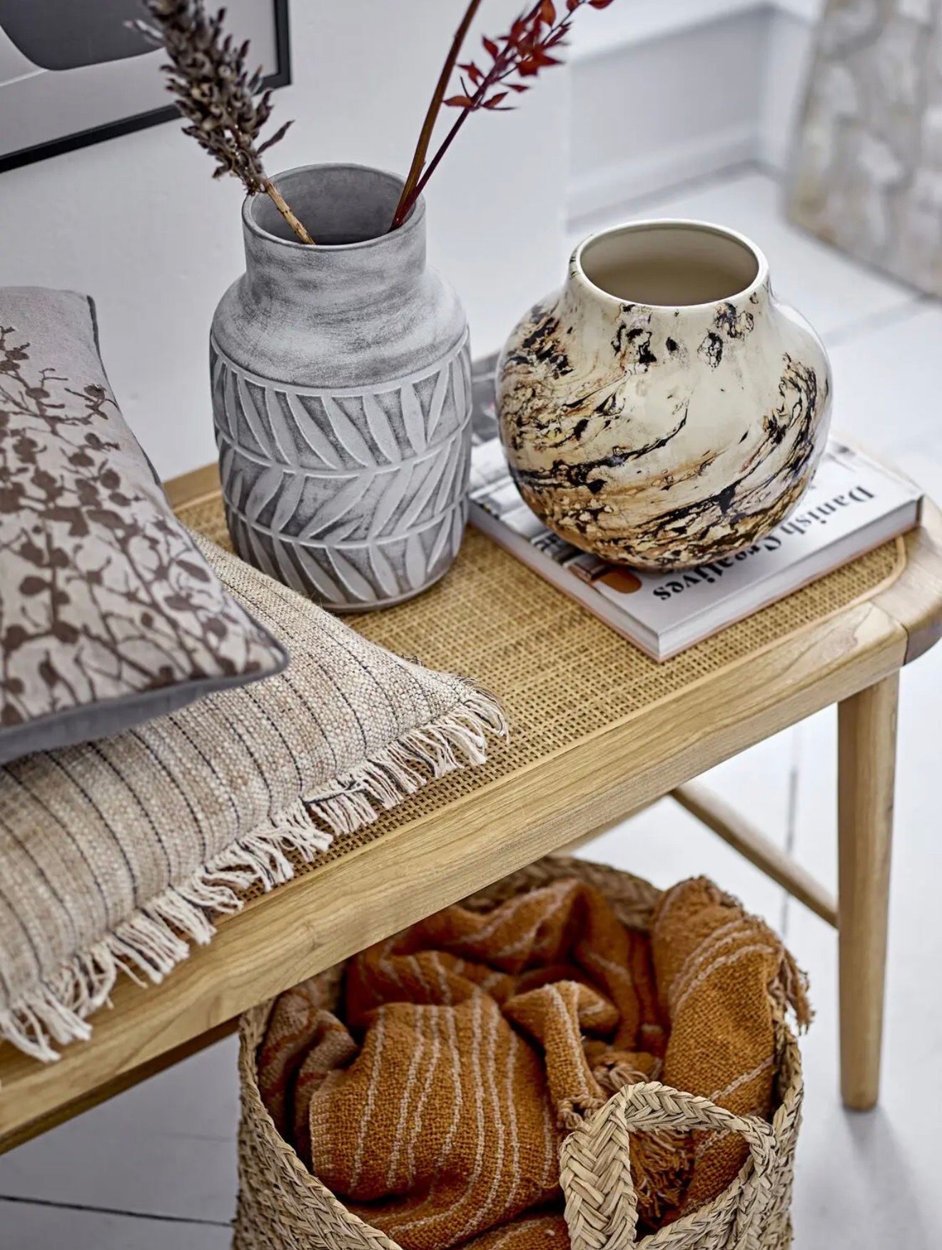 A light wood bench with a woven top displays a grey textured vase with dried branches, a marbled vase, a book, and two pillows. A woven basket with an orange blanket is underneath.