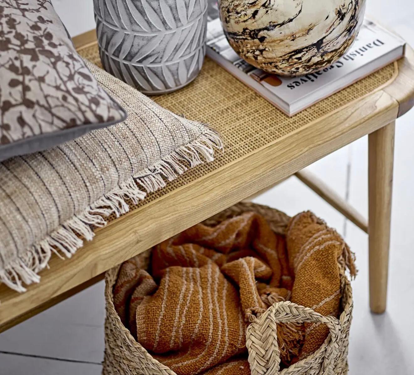 A light wood bench with a woven top displays a grey textured vase with dried branches, a marbled vase, a book, and two pillows. A woven basket with an orange blanket is underneath.