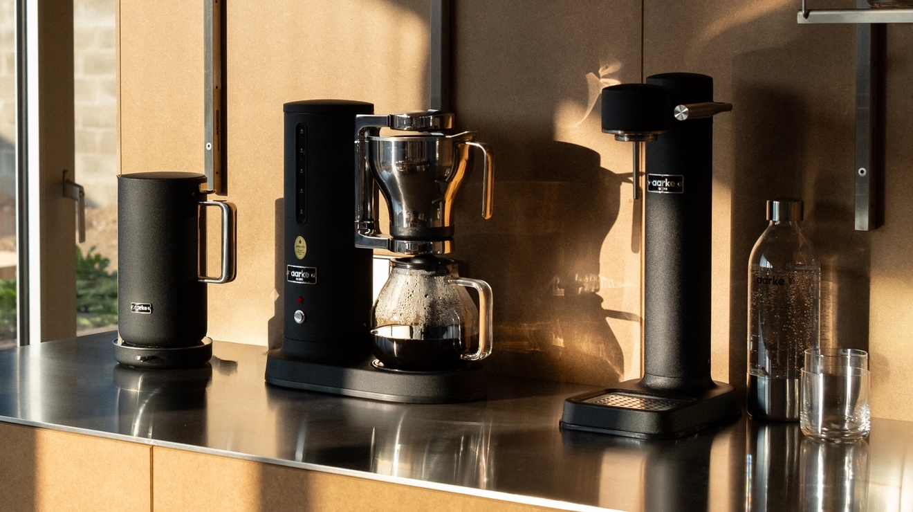 Svart kaffekoker, dryppmaskin og kvern på en metallbenk, ved siden av et vindu med utsikt over en hage.