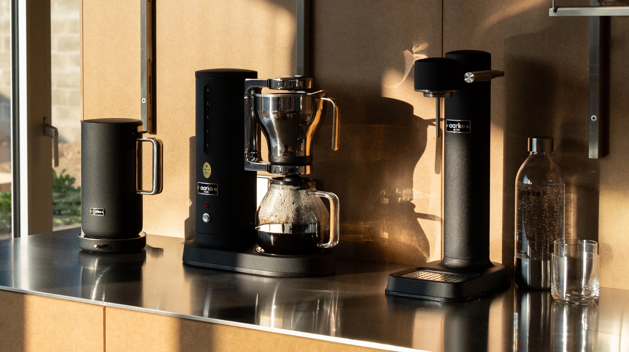 Svart kaffekoker, dryppmaskin og kvern på en metallbenk, ved siden av et vindu med utsikt over en hage.
