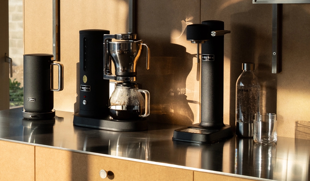 Svart kaffekoker, dryppmaskin og kvern på en metallbenk, ved siden av et vindu med utsikt over en hage.