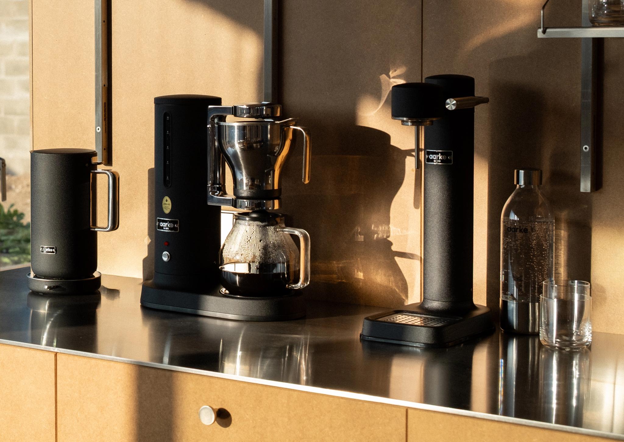 Svart kaffemaskin och svart vattenkokare från Aarke på en modern köksbänk med solig trädgårdsutsikt.