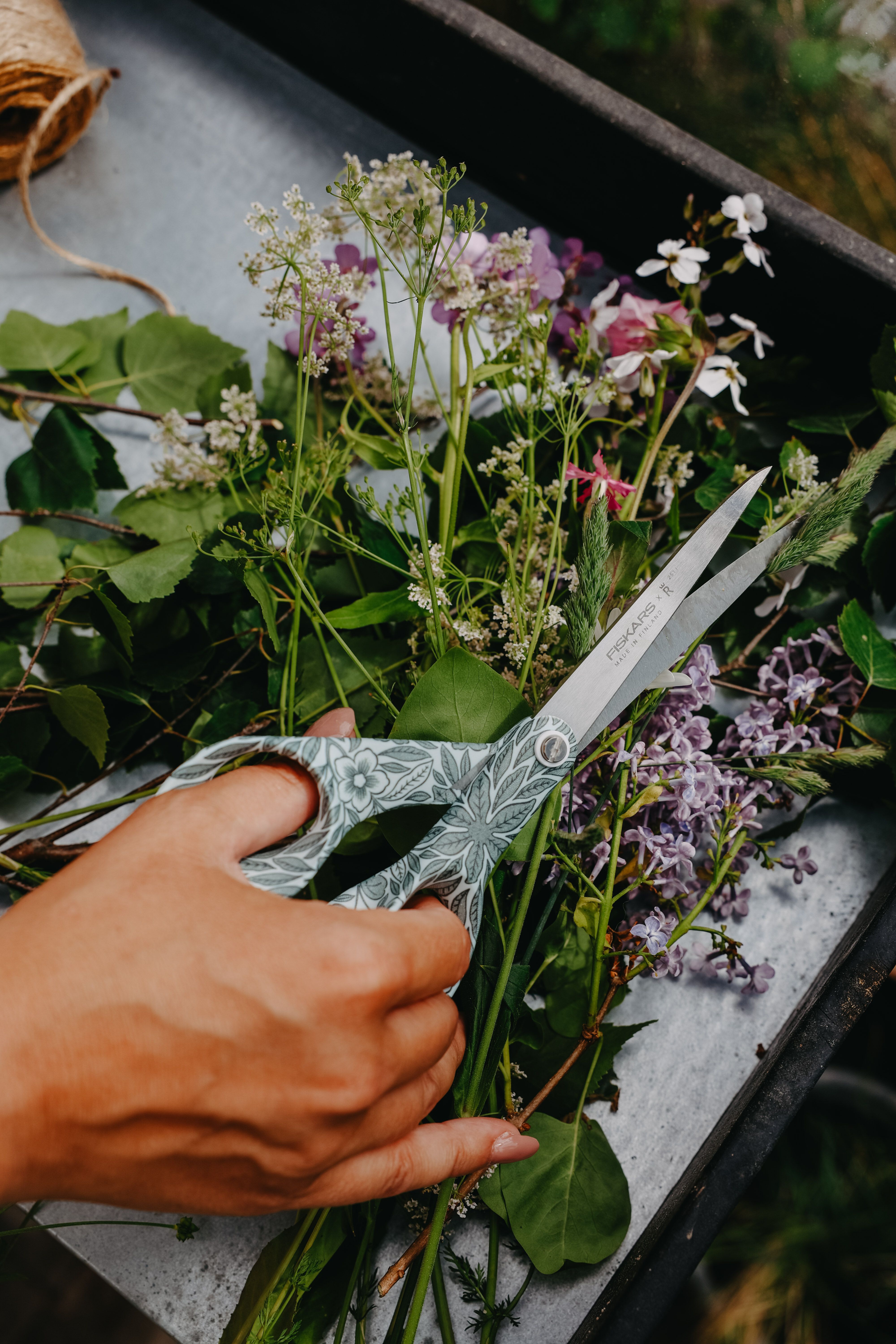 Hand som klipper nyplockade blommor och gröna kvistar med ett mönstrat trädgårdssax på ett bord.