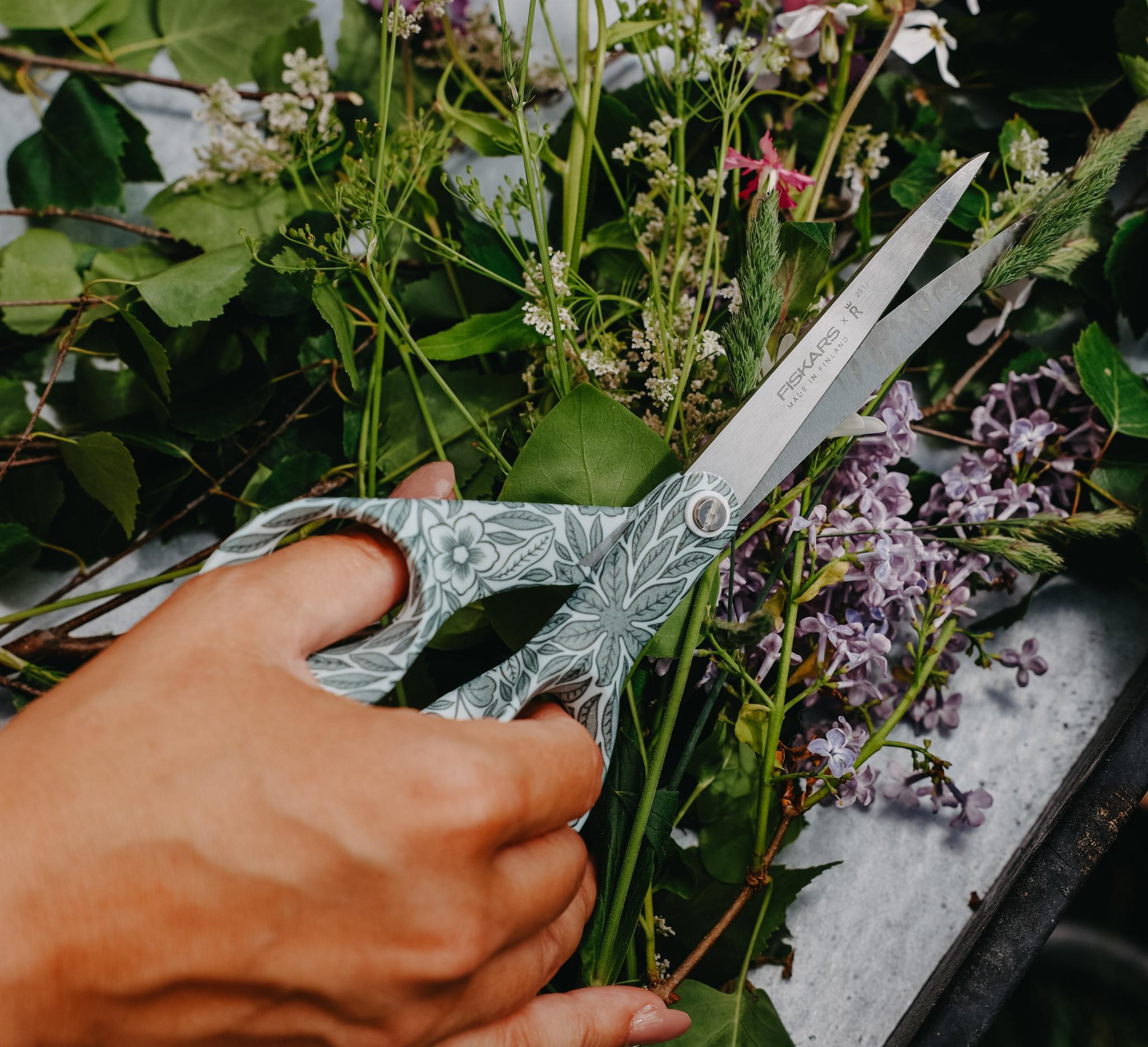 Hand som klipper nyplockade blommor och gröna kvistar med ett mönstrat trädgårdssax på ett bord.