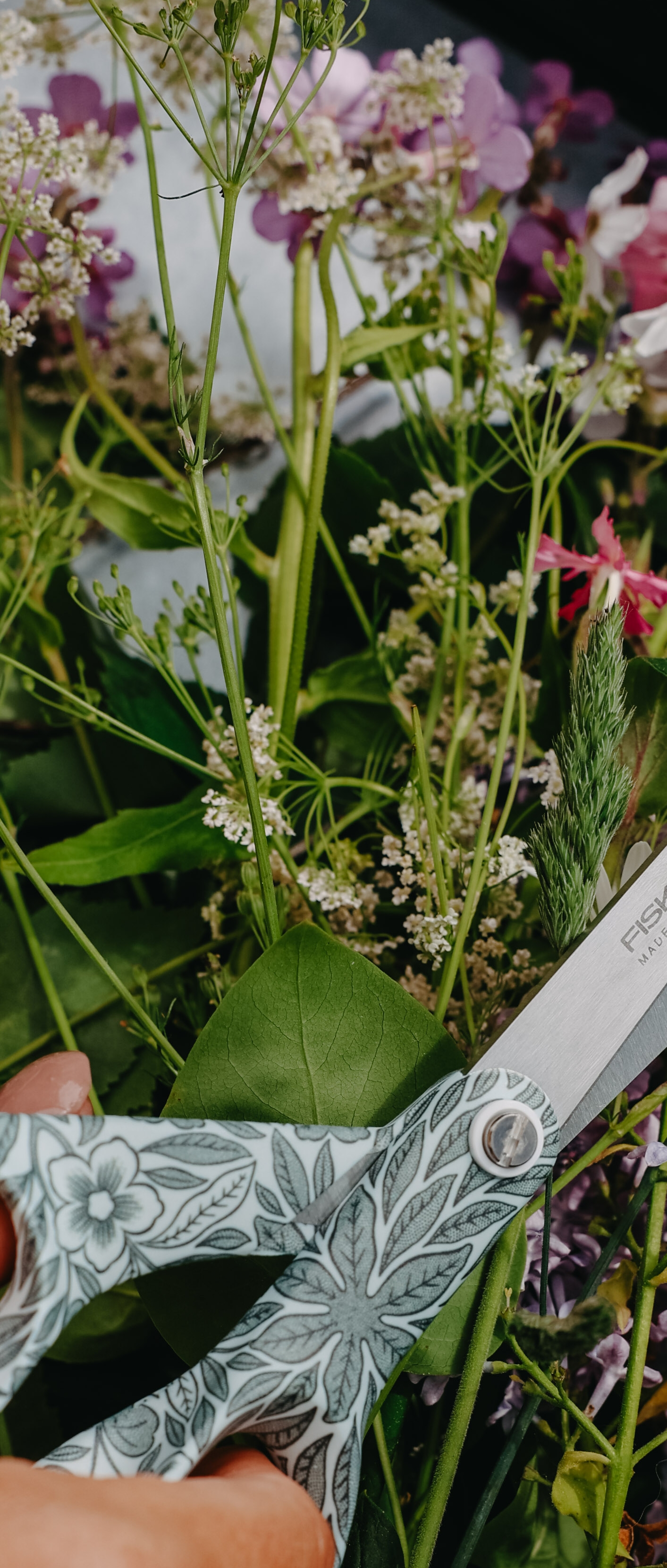 Hand som klipper nyplockade blommor och gröna kvistar med ett mönstrat trädgårdssax på ett bord.