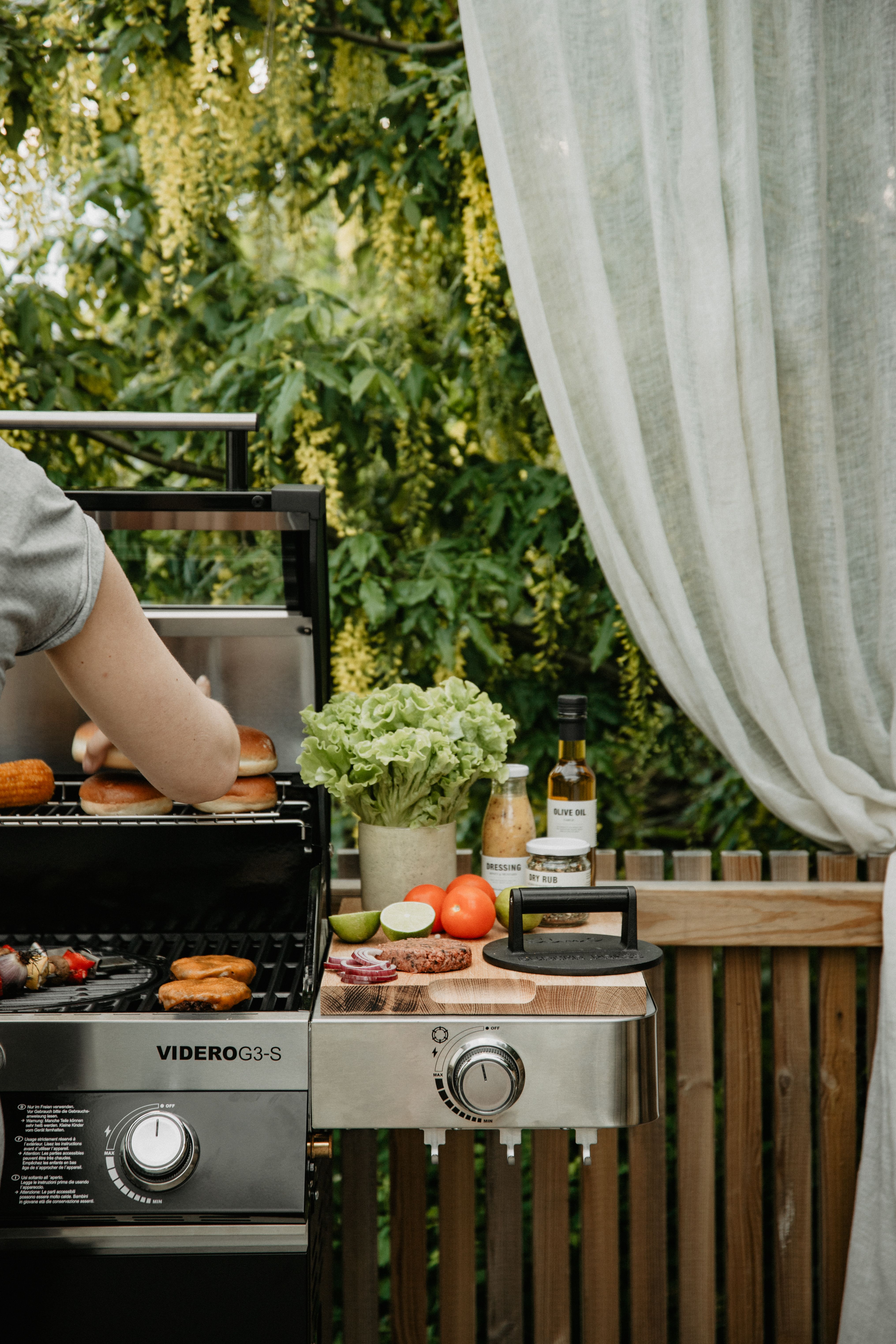 En person grillar hamburgare, bröd och majs på en gasolgrill utomhus, med färska ingredienser och tillbehör på ett sidobord