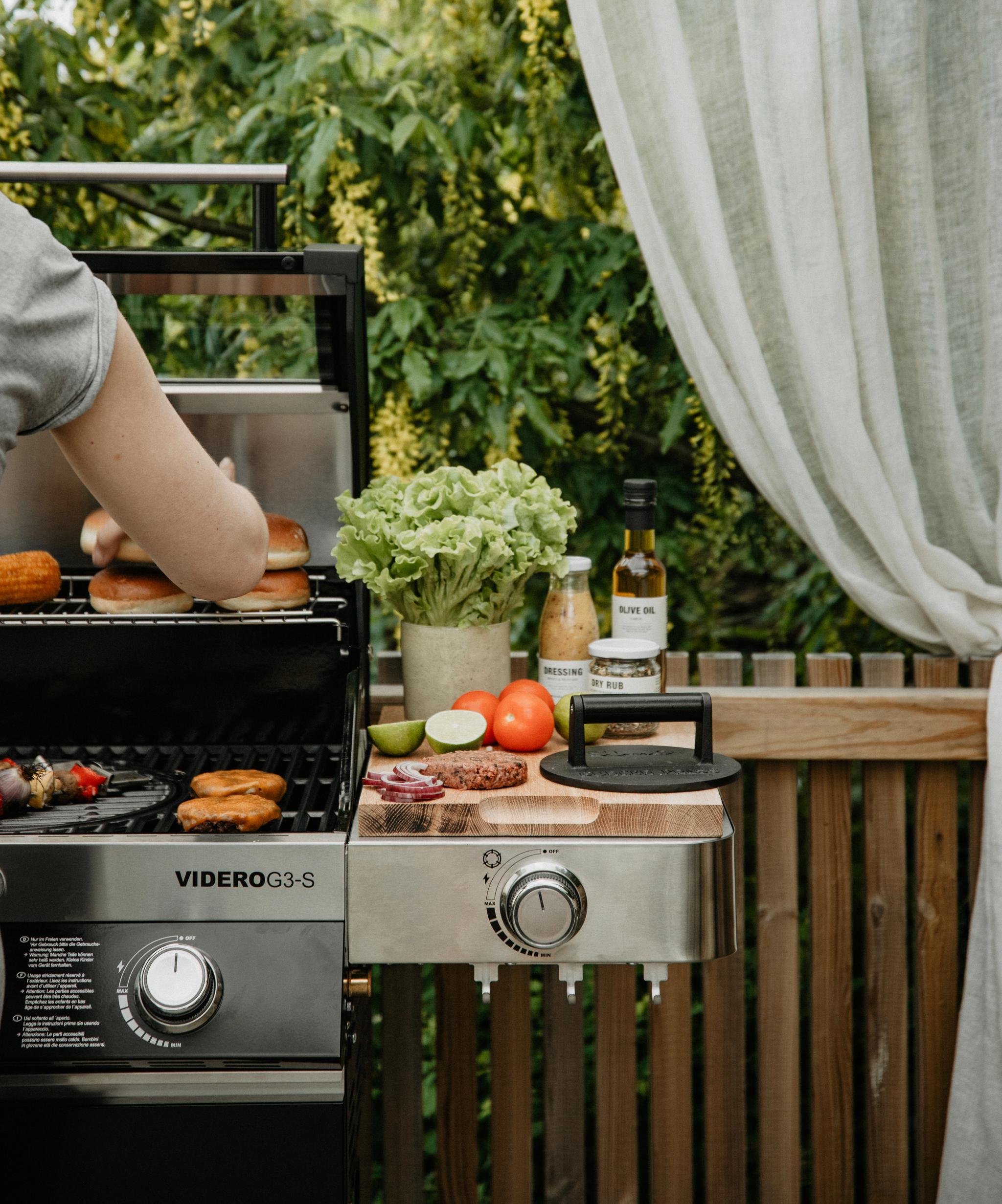 En person griller burgere, brød og mais på en utendørs gassgrill, med ferske ingredienser og tilbehør på et sidebord