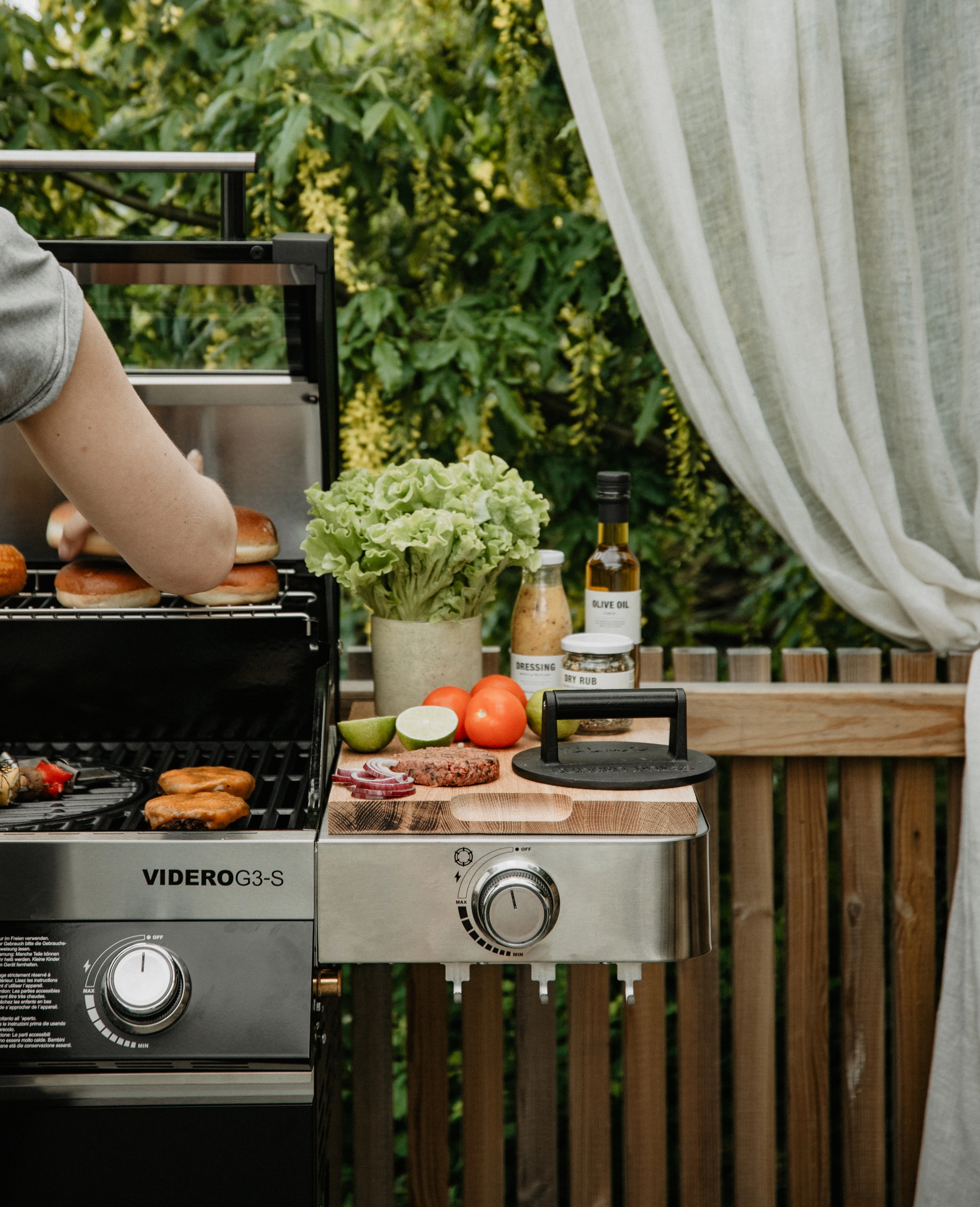 En person grillar hamburgare, bröd och majs på en gasolgrill utomhus, med färska ingredienser och tillbehör på ett sidobord