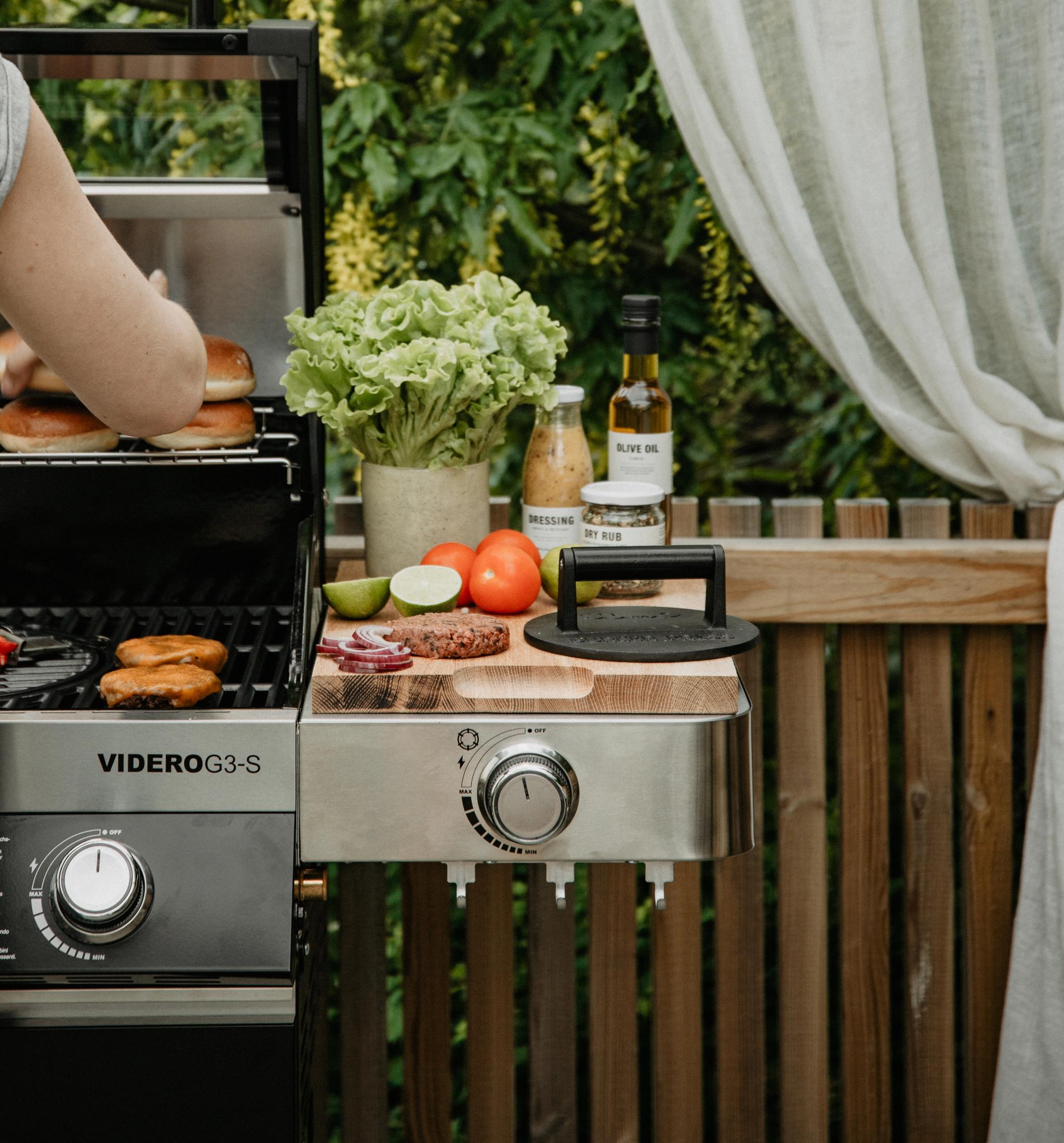 En person grillar hamburgare, bröd och majs på en gasolgrill utomhus, med färska ingredienser och tillbehör på ett sidobord