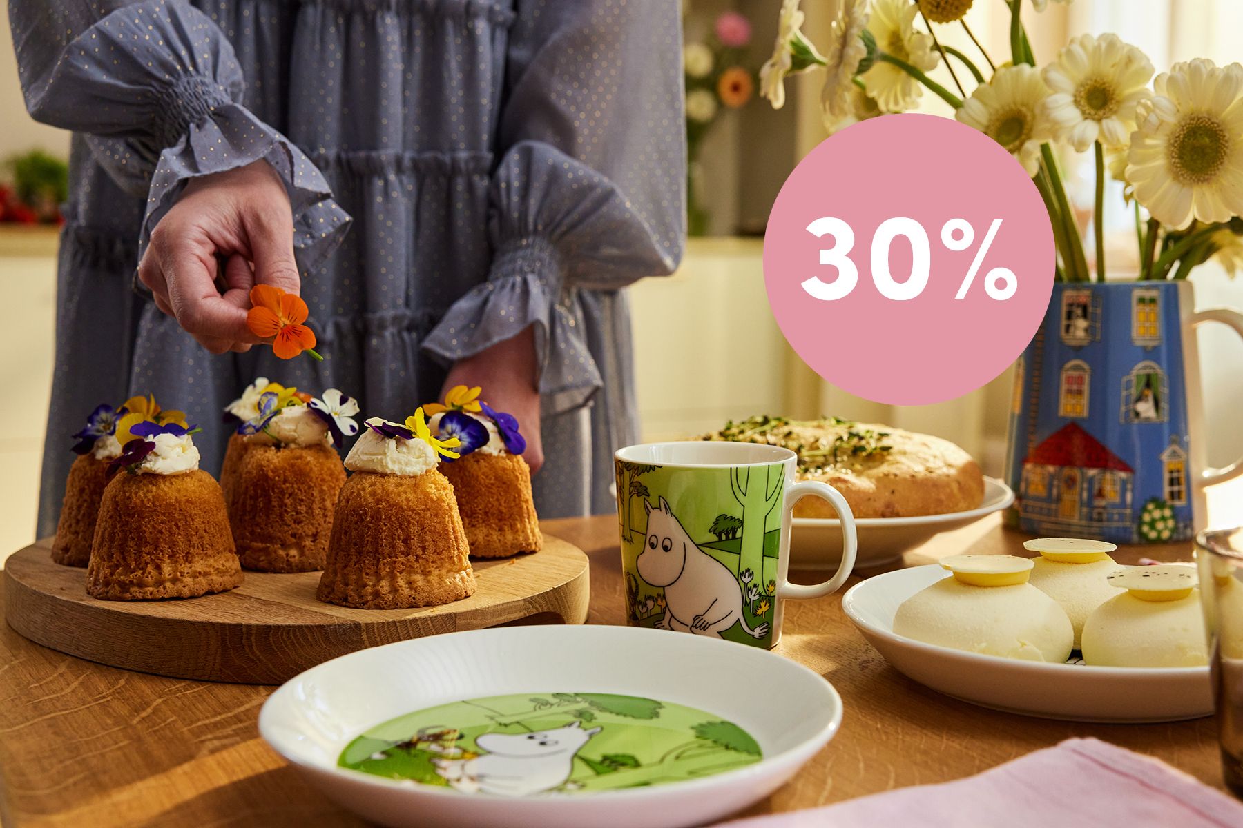 En person pynter små kaker med blomster på et bord dekket med Mummi-Arabia-skåler og en grafikk med «30 % rabatt på Mummi-Arabia»-salg.